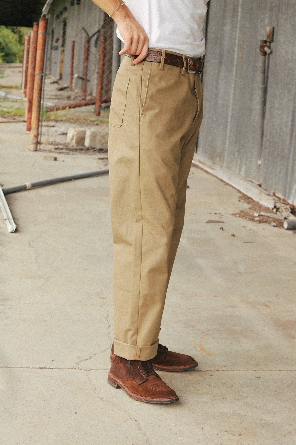 A person wearing khaki chino pants, a white shirt, and brown leather boots standing outdoors against a corrugated metal wall.