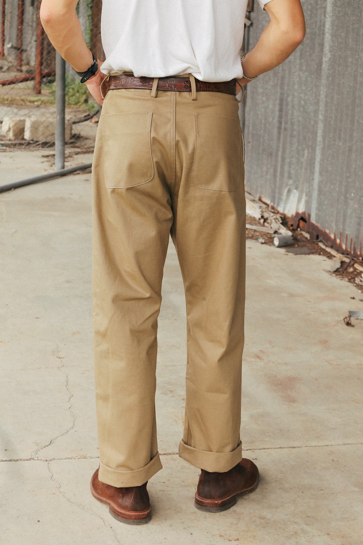 A person shown from the back wearing tan chinos with a white t-shirt, brown belt, and brown leather shoes, standing on a concrete surface.