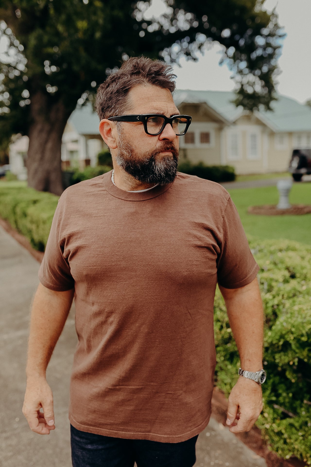 Man with beard and glasses wearing a brown t-shirt standing outdoors near green bushes and houses