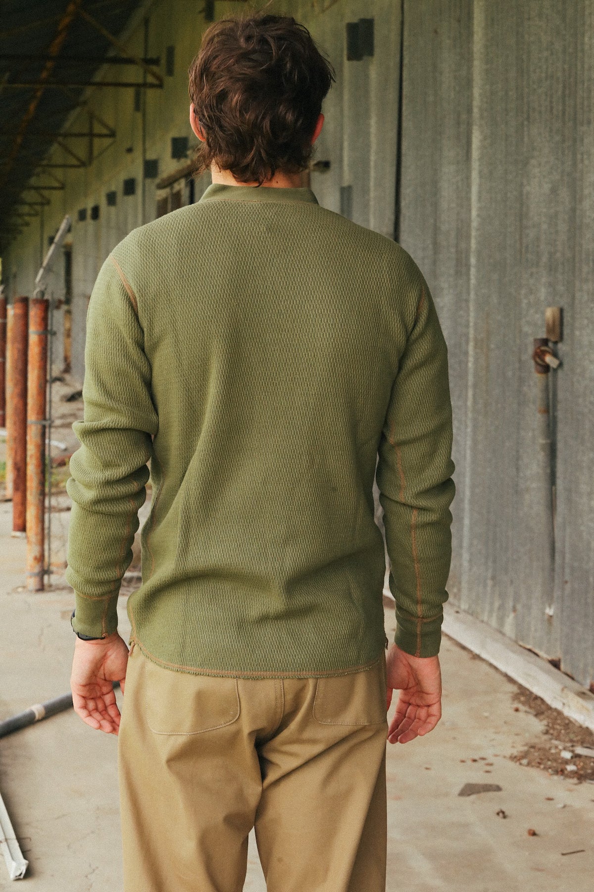 A man viewed from behind wearing an olive green ribbed knit sweater and tan pants, standing in a rustic industrial corridor with corrugated metal walls.