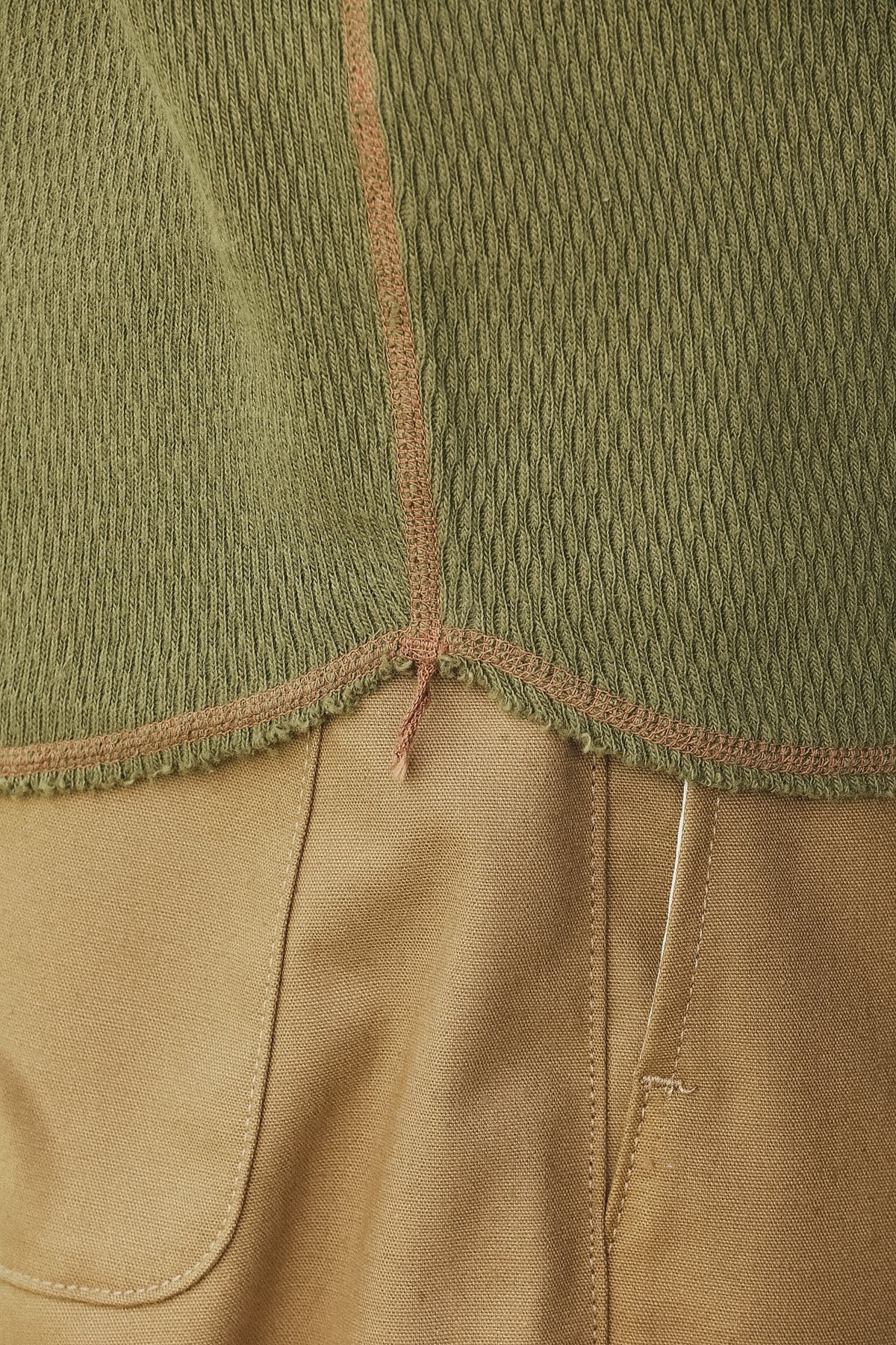 Close-up detail of a garment showing an olive green textured knit upper section with a frayed hem and a tan lower section with visible stitching and pocket seams.