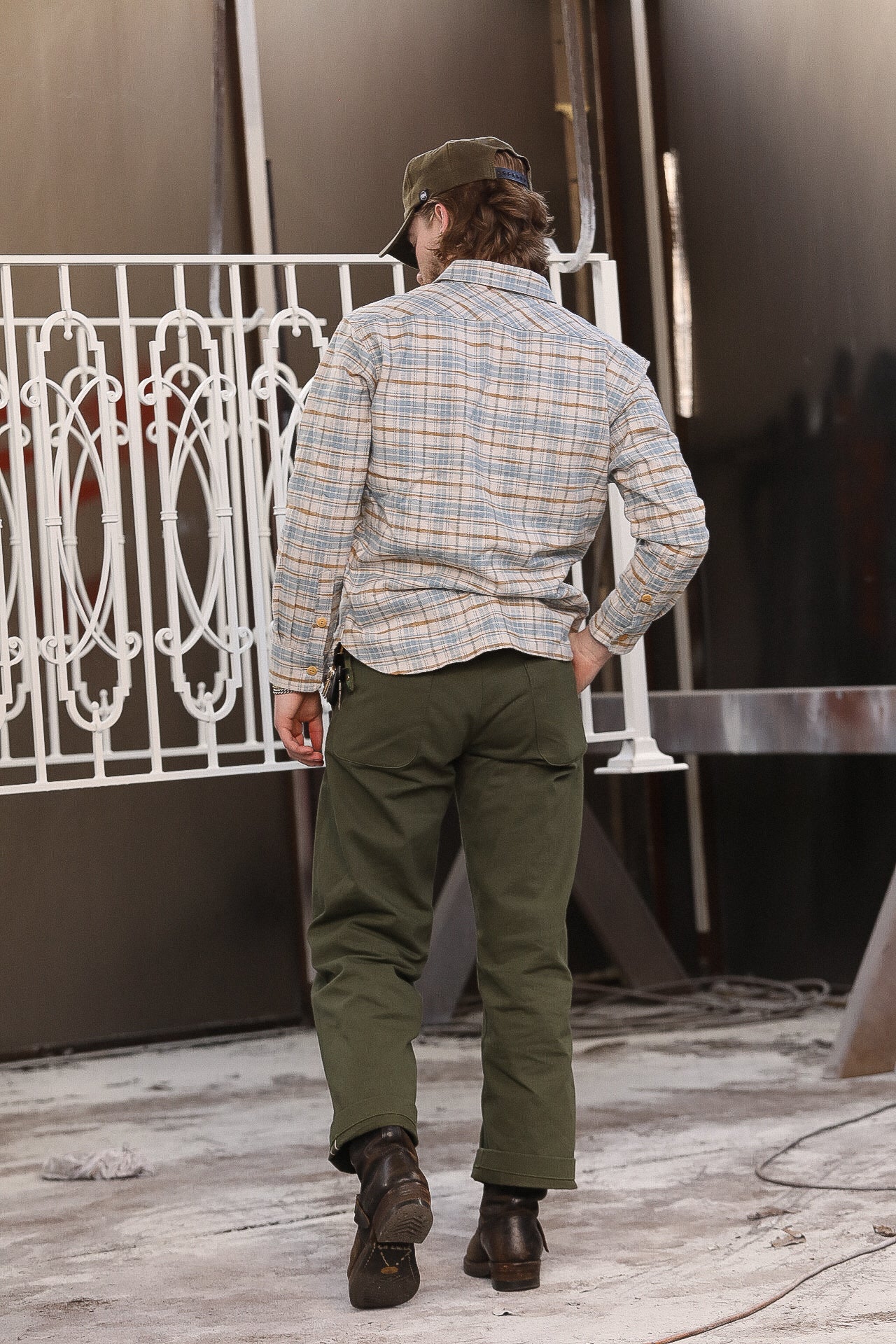 Man in plaid shirt, olive pants, and boots standing in industrial workshop with metal railing
