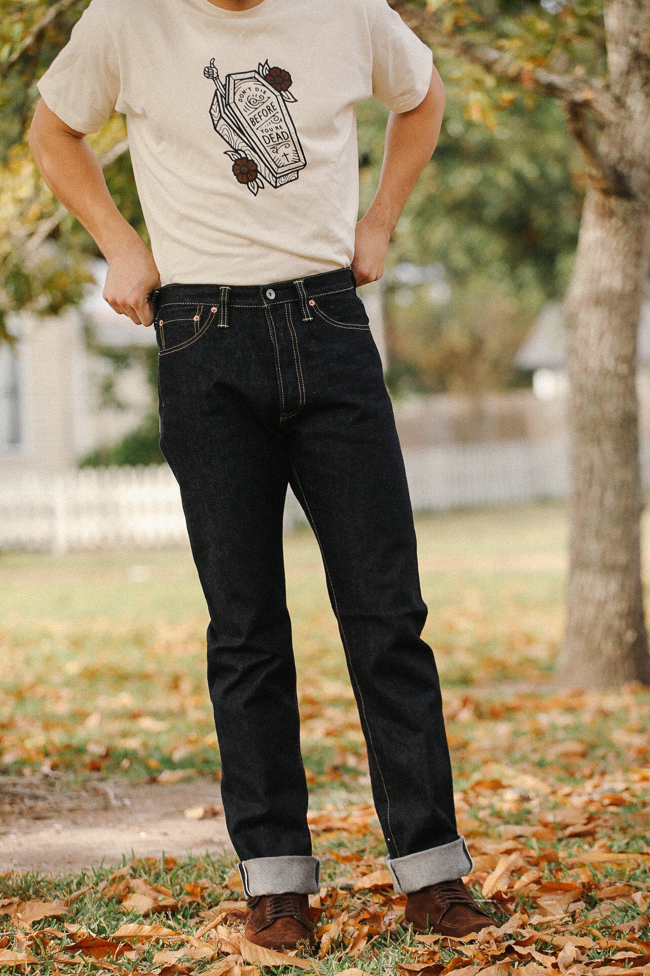 Man wearing black raw denim jeans with rolled cuffs and brown shoes, standing on fallen leaves outdoors
