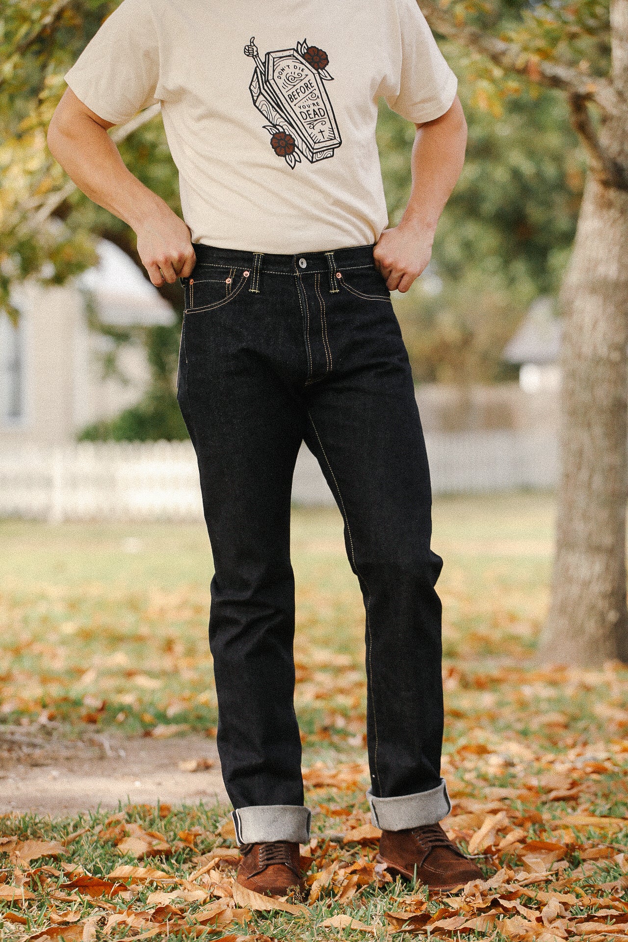 Man wearing dark raw denim jeans with cuffed hems and brown leather shoes standing outdoors in autumn