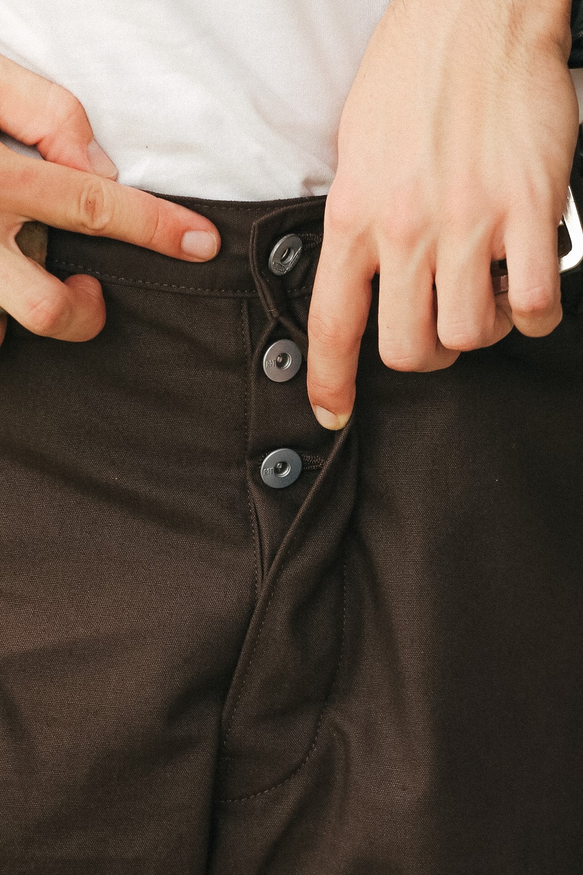 Close-up of a person wearing dark brown pants with a white shirt, showing hands adjusting the waistband and pointing to the three front buttons.