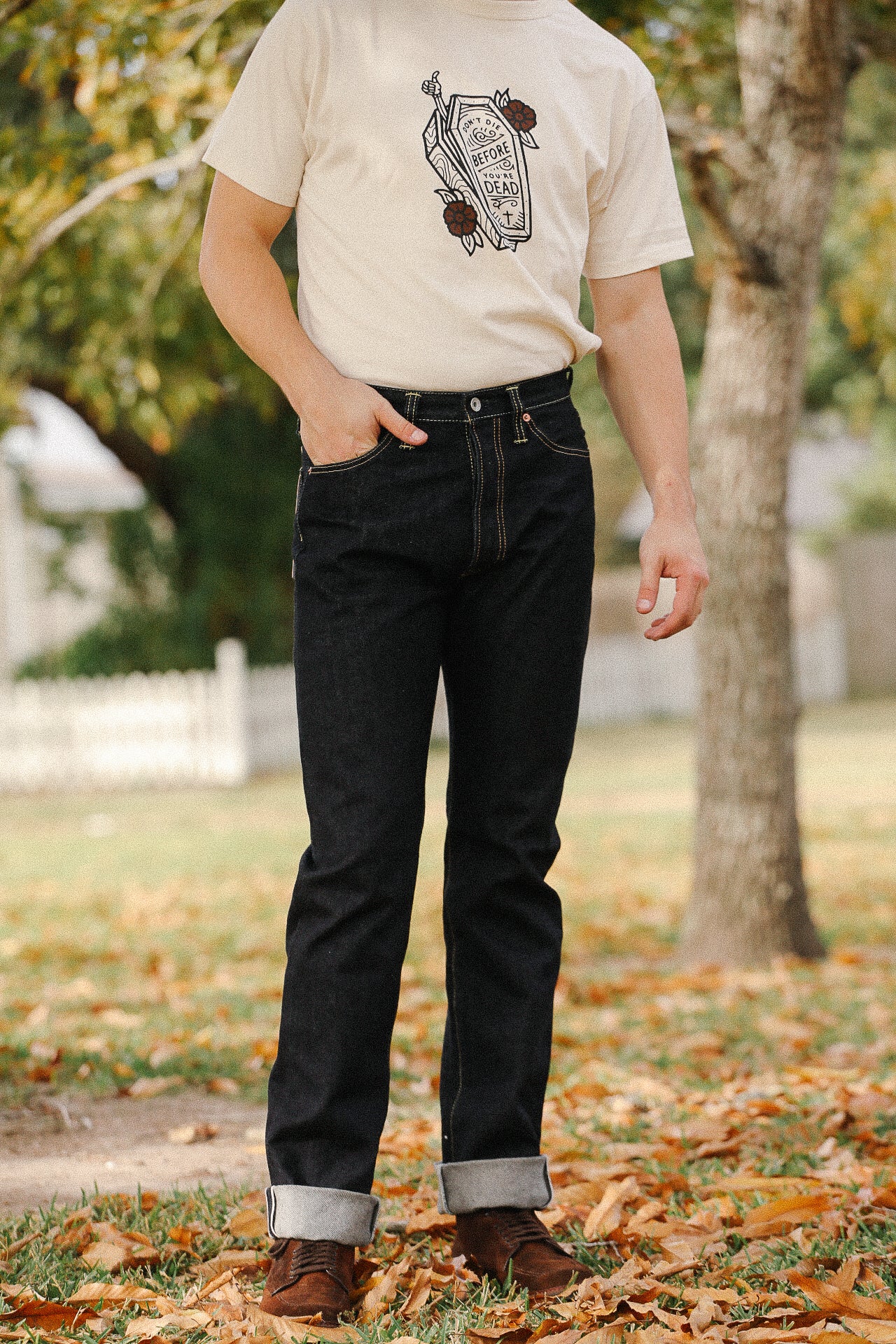 Man wearing dark raw denim jeans with cuffs and brown leather shoes, paired with a graphic tee outdoors