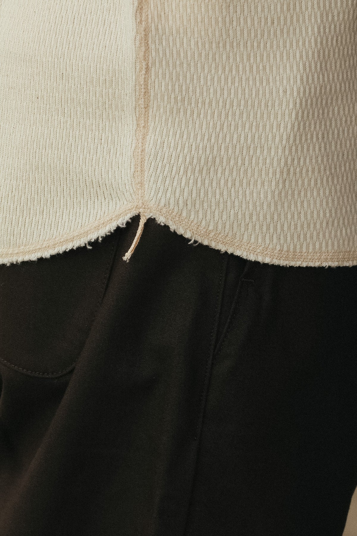 Close-up detail of a cream-colored textured knit top with a raw, frayed hem, paired with black trousers.