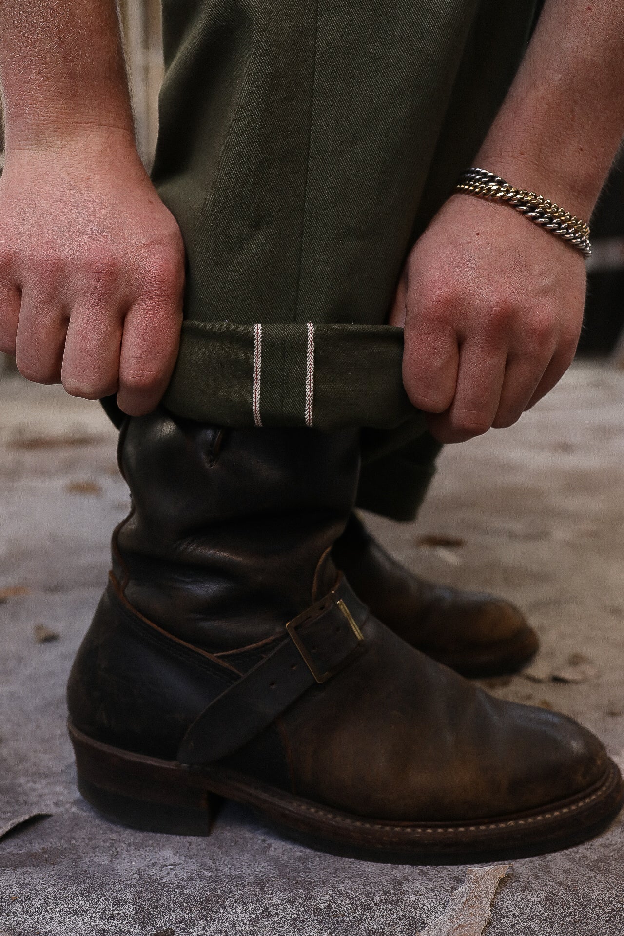 Close-up of hands rolling up olive green selvedge denim over worn brown leather engineer boots