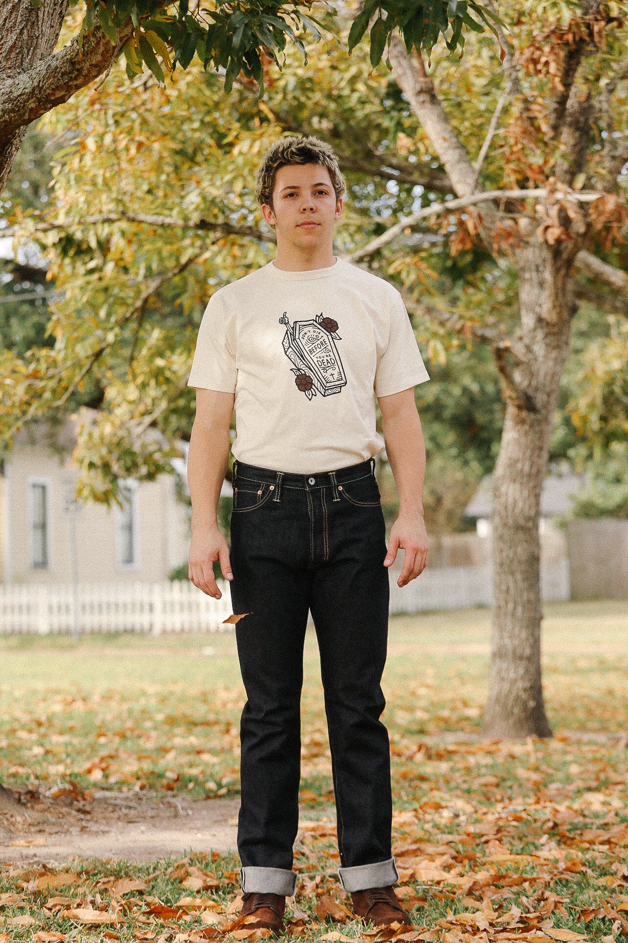 Man wearing light t-shirt with coffin graphic and dark rolled-up jeans standing outdoors among autumn leaves