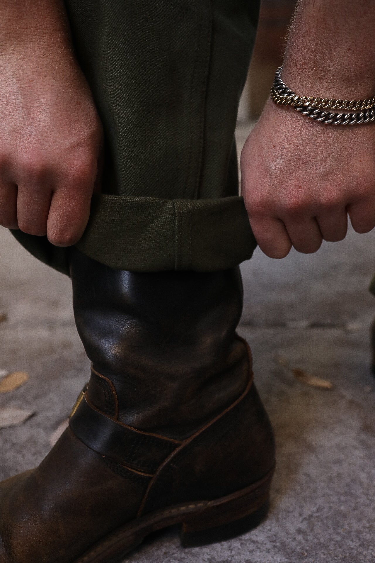 Close-up of hands rolling up olive green pants above worn brown leather boots with a silver and gold chain bracelet