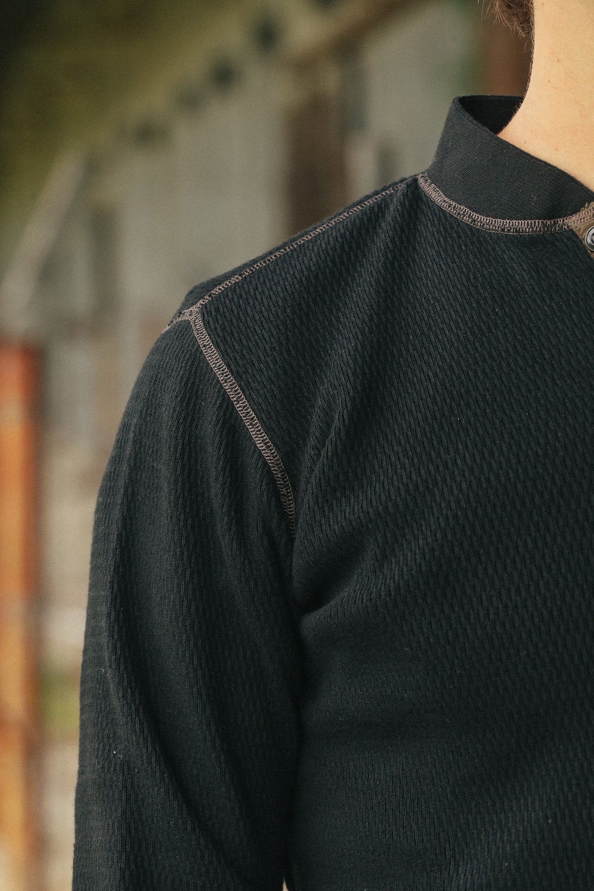 Close-up of a person wearing a dark textured long-sleeve top with a stand collar and contrast stitching, photographed outdoors with a blurred background.