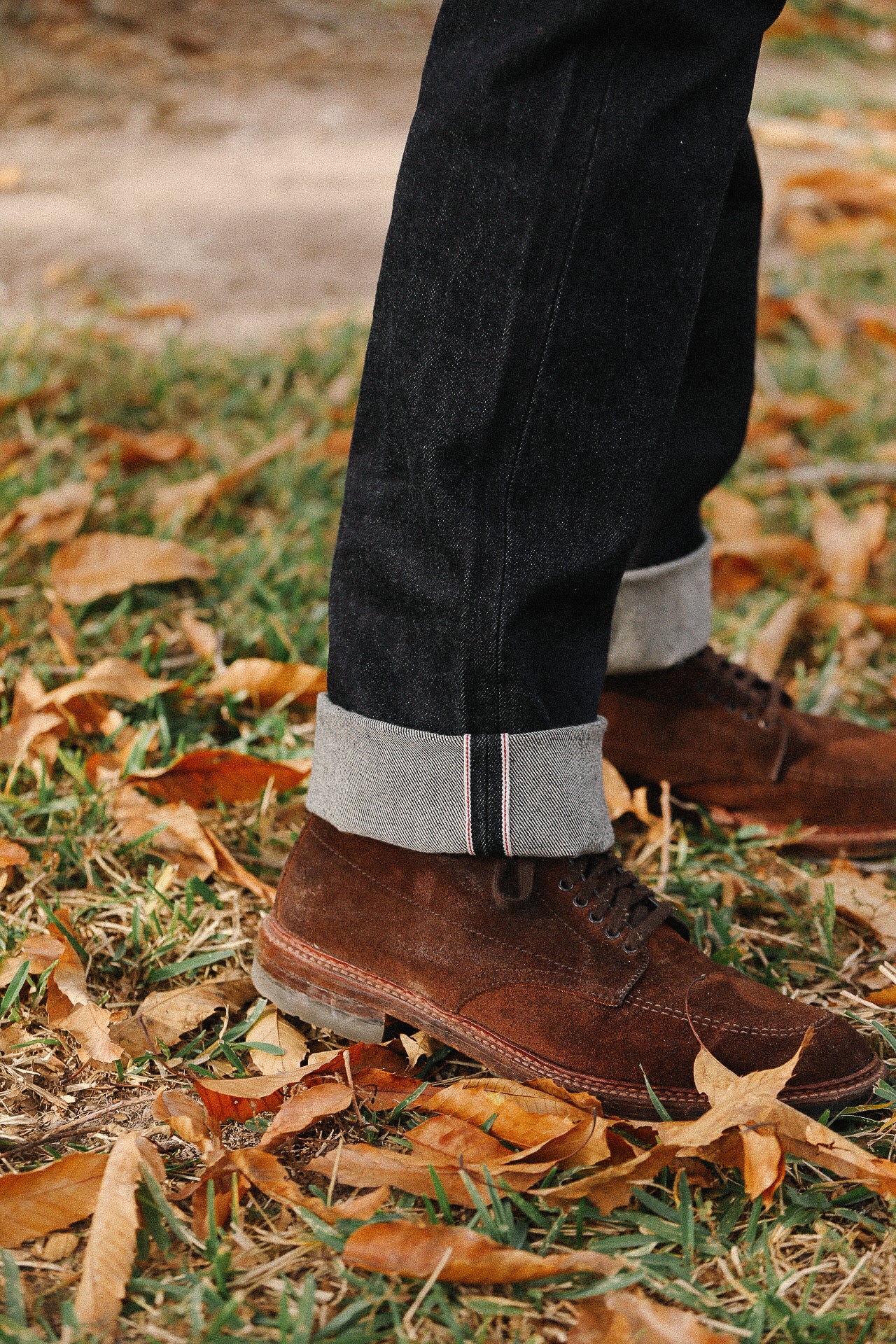 Close-up of brown suede boots paired with dark selvedge denim jeans cuffed above autumn leaves