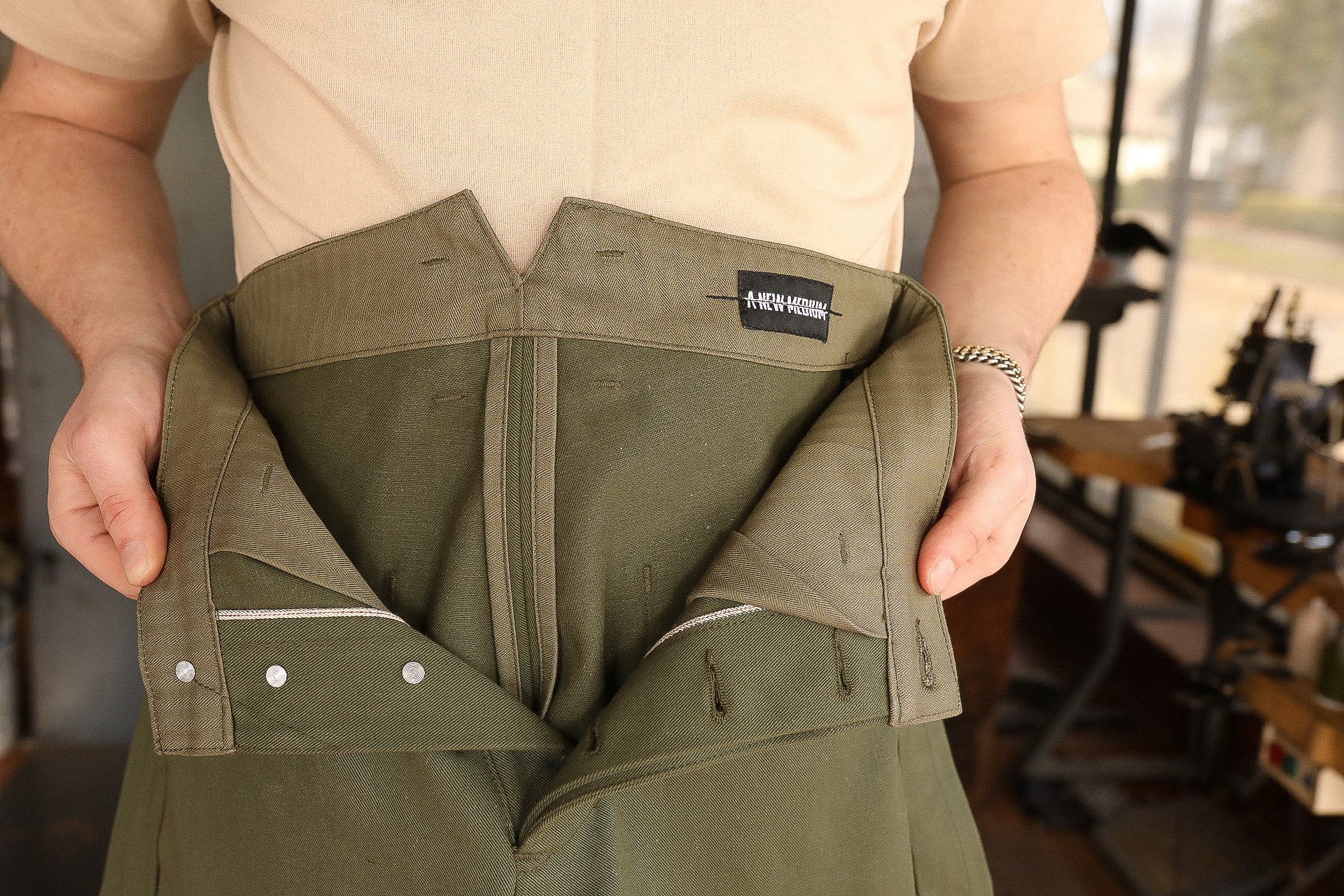 Person holding open a pair of olive green work pants with detailed internal stitching and label in workshop