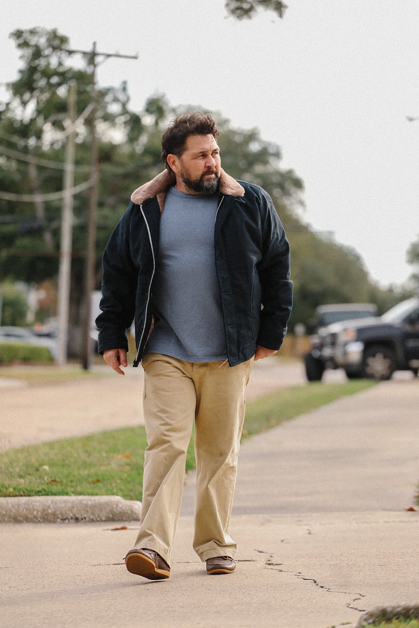 Man wearing black jacket with fur collar, blue shirt, khaki pants walking on sidewalk in casual outdoor setting
