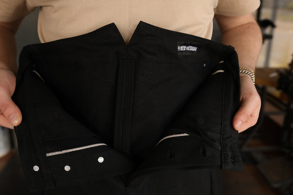 Hand holding open black selvedge denim jeans showing buttons and interior stitching detail
