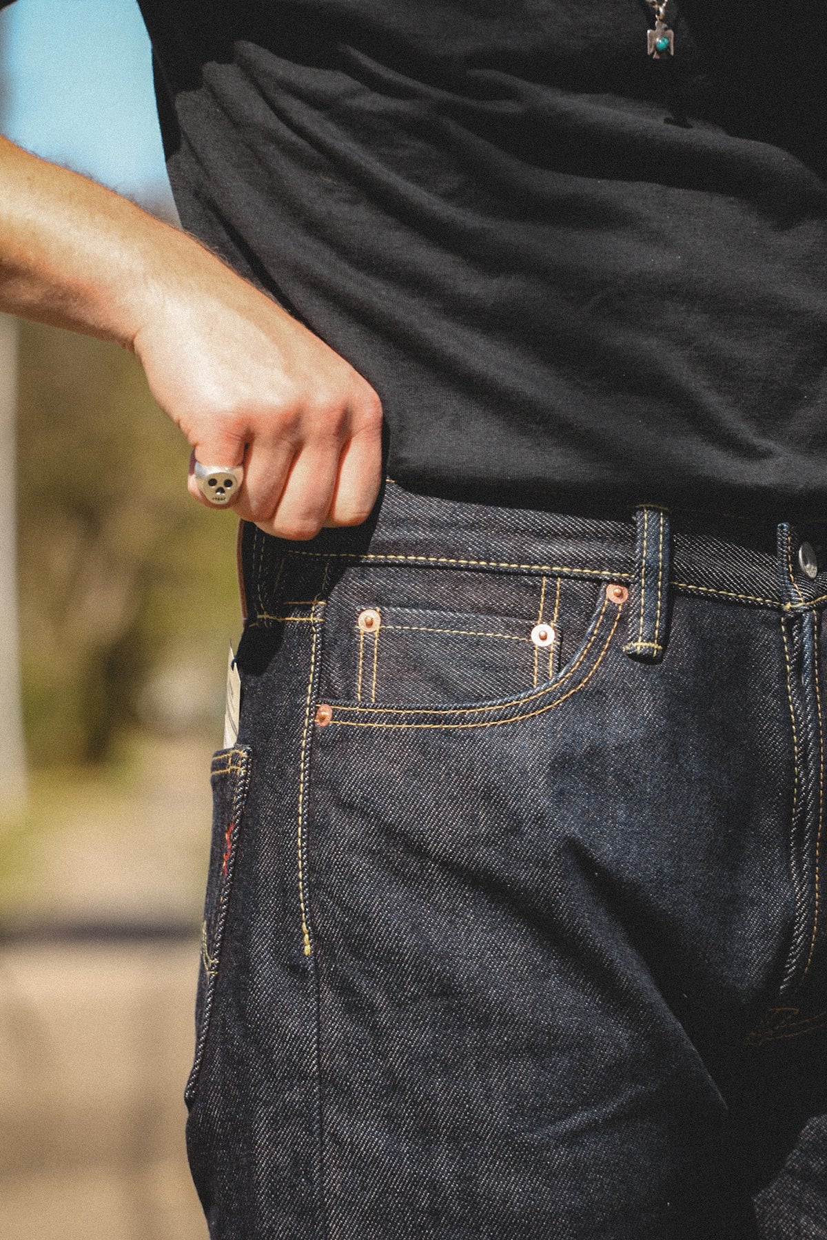 Close-up of a person wearing dark indigo denim jeans with contrast stitching and a black t-shirt, pulling the shirt slightly away from the waistband.