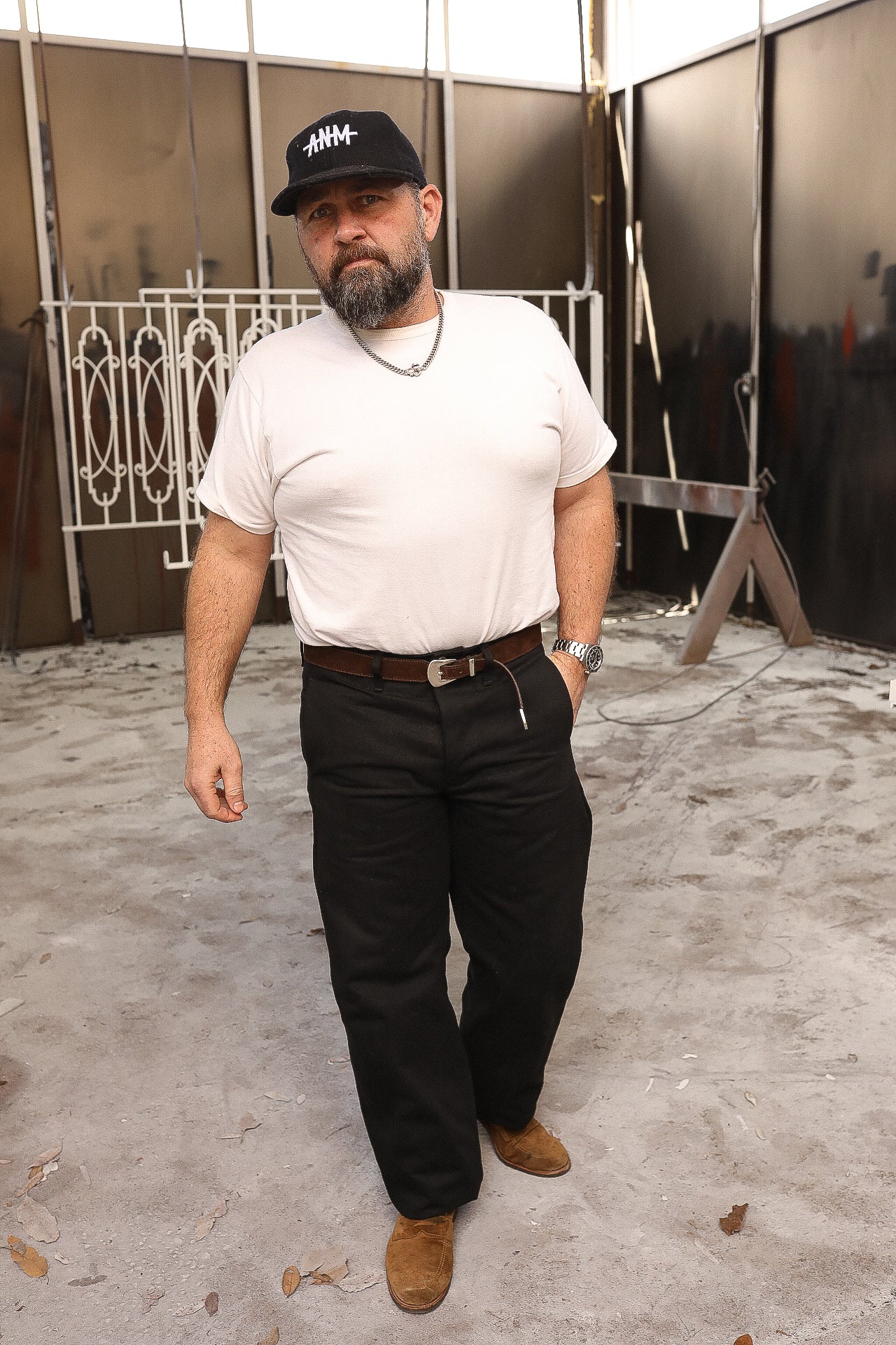 Bearded man wearing white t-shirt, black pants, and black cap standing in industrial metal workshop