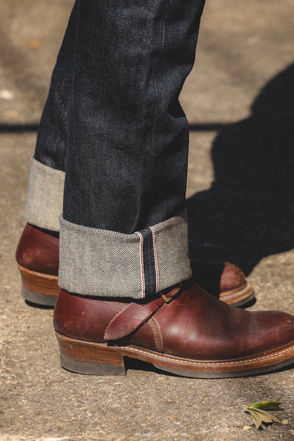 A close-up of a person's legs wearing dark cuffed denim jeans and brown leather boots on a concrete surface.