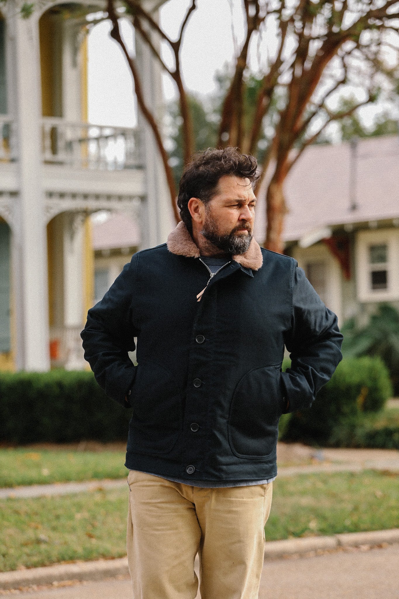 Man wearing black jacket with brown shearling collar and beige pants standing outdoors in front of houses