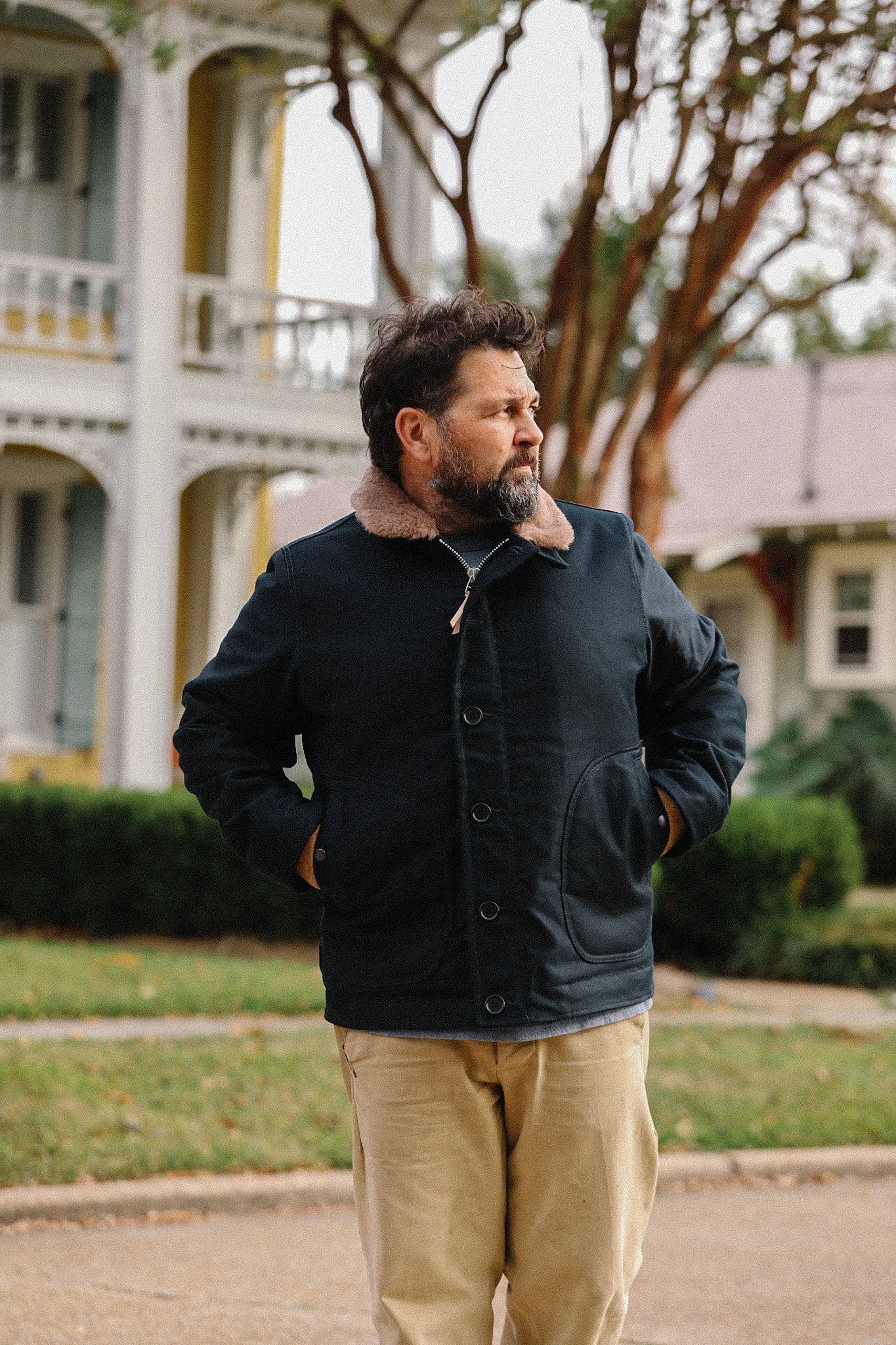 Man wearing black shearling collar jacket and khaki pants outdoors near vintage house