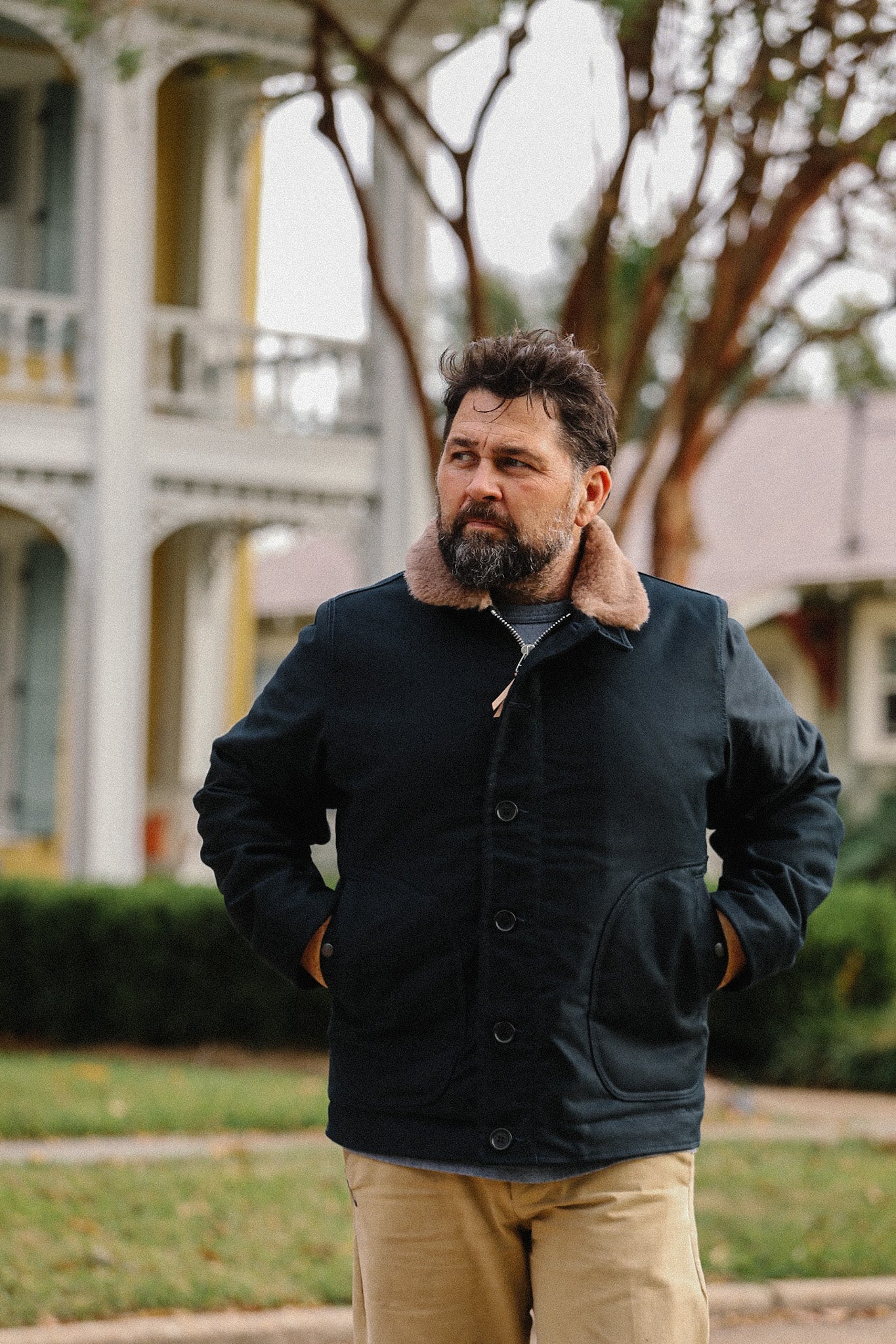 Bearded man wearing a dark navy jacket with a fur collar and tan pants standing outdoors in front of a white house