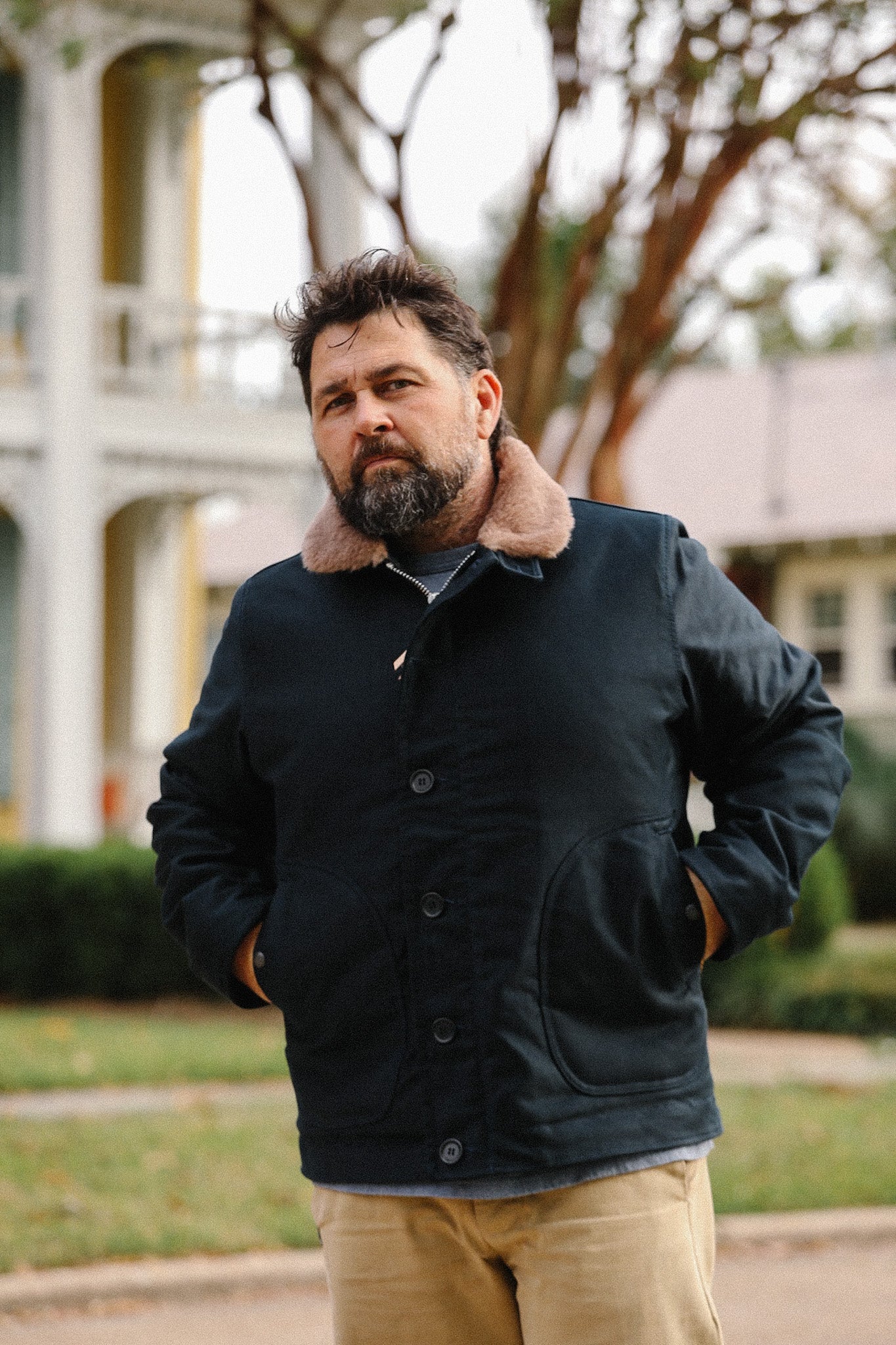 Bearded man wearing a black jacket with fur collar outdoors near white colonial-style building