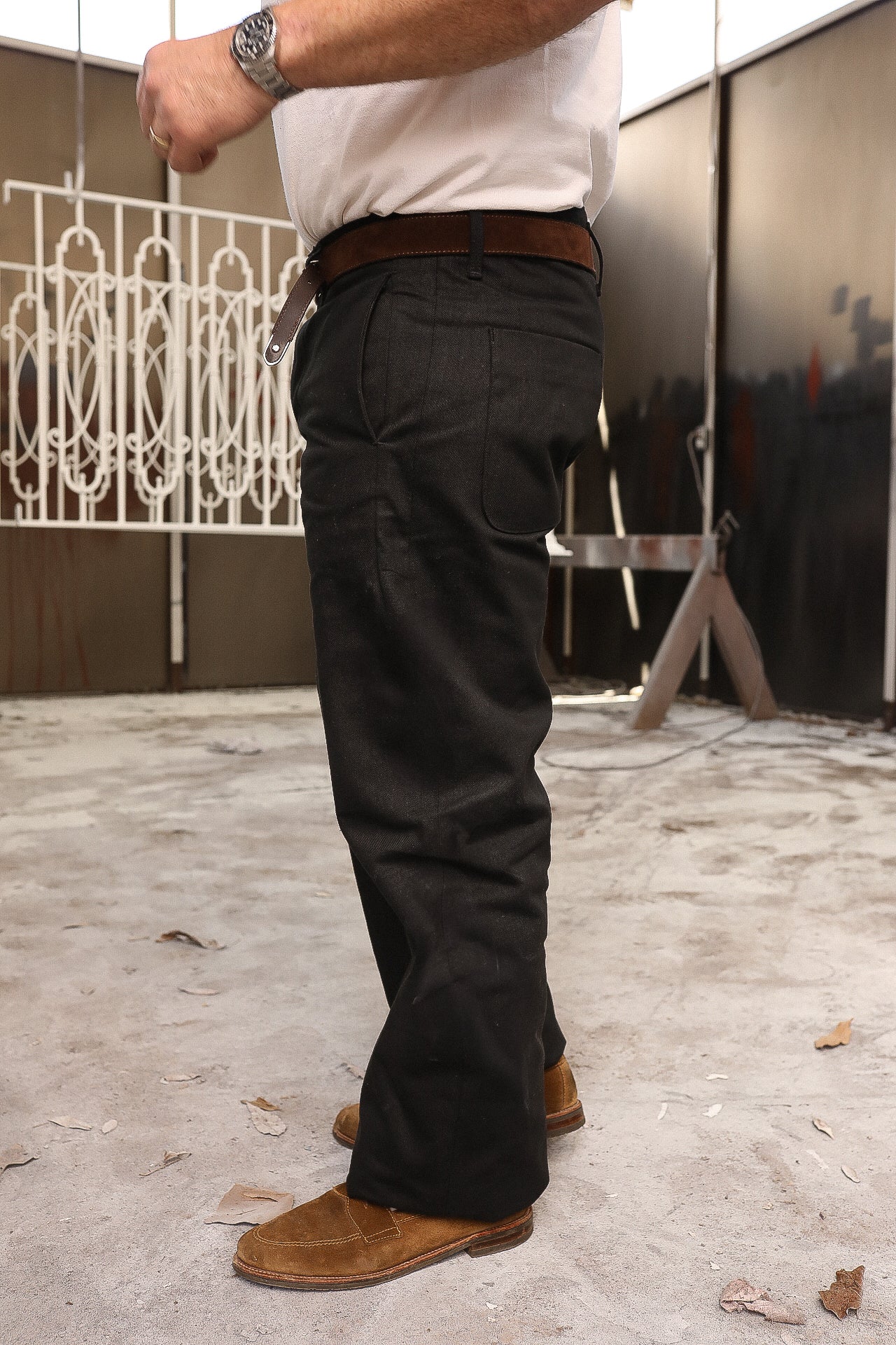 Man wearing black work pants with a brown leather belt and brown suede boots in a workshop setting
