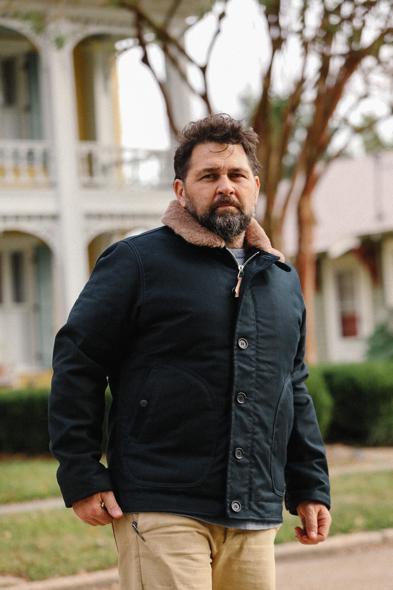 Man wearing black jacket with brown shearling collar outdoor in front of white building