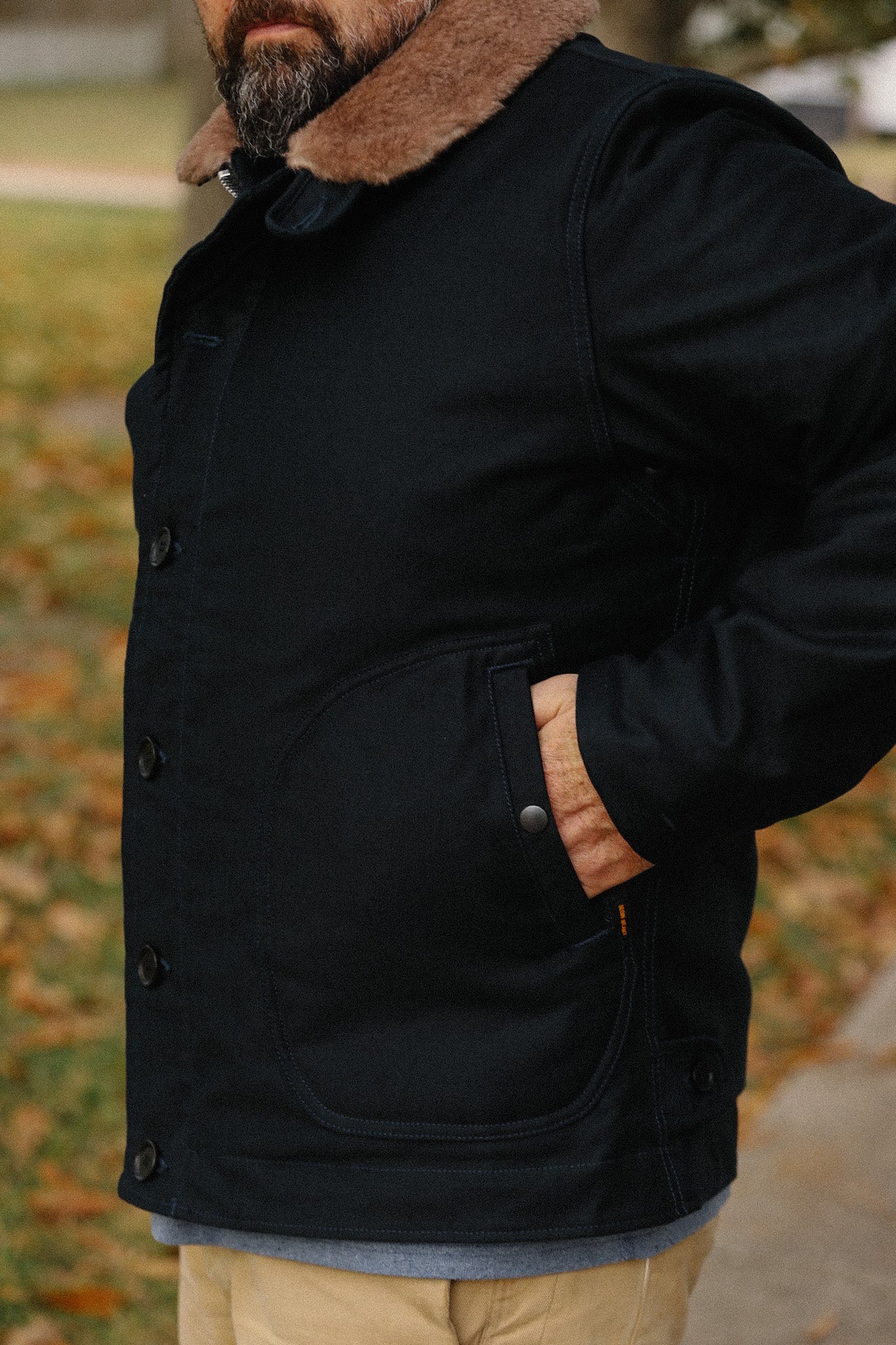 Man wearing black work jacket with brown fur collar and hand in pocket outdoors on autumn day