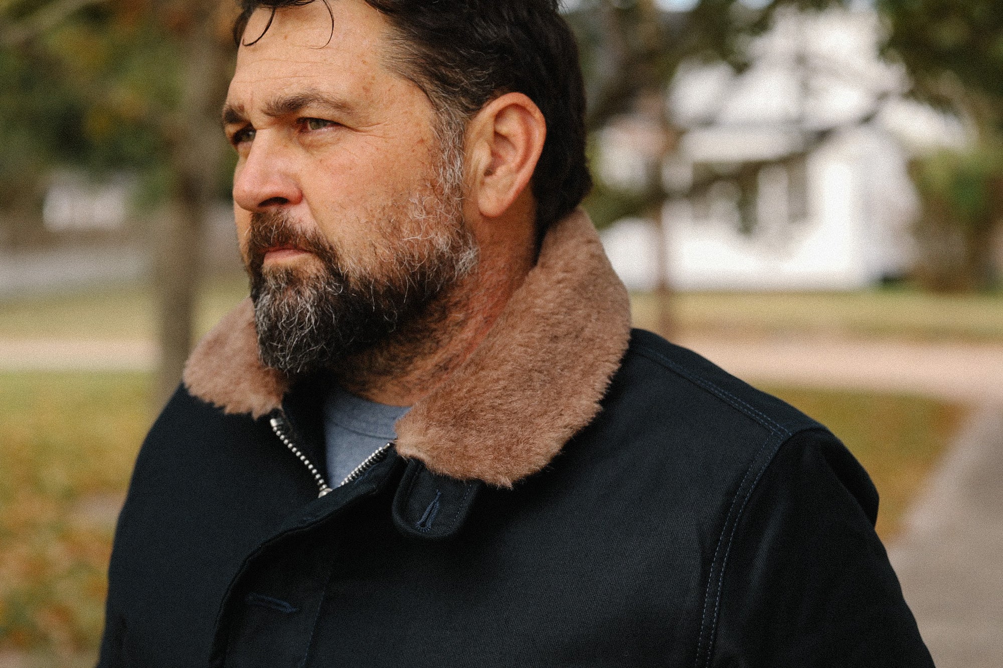 Man with beard wearing black jacket with tan shearling collar outdoors in fall scenery