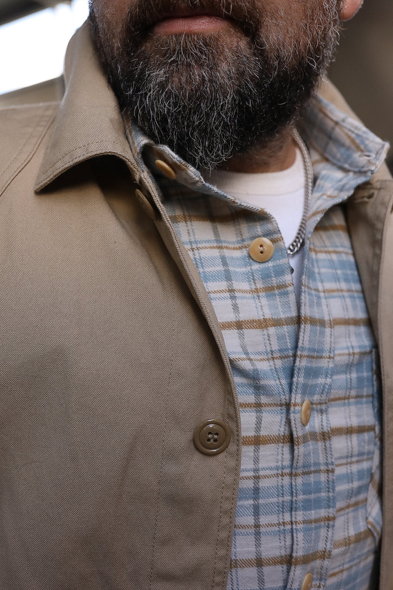 Close-up of a bearded man wearing a beige jacket over a blue and brown plaid flannel shirt