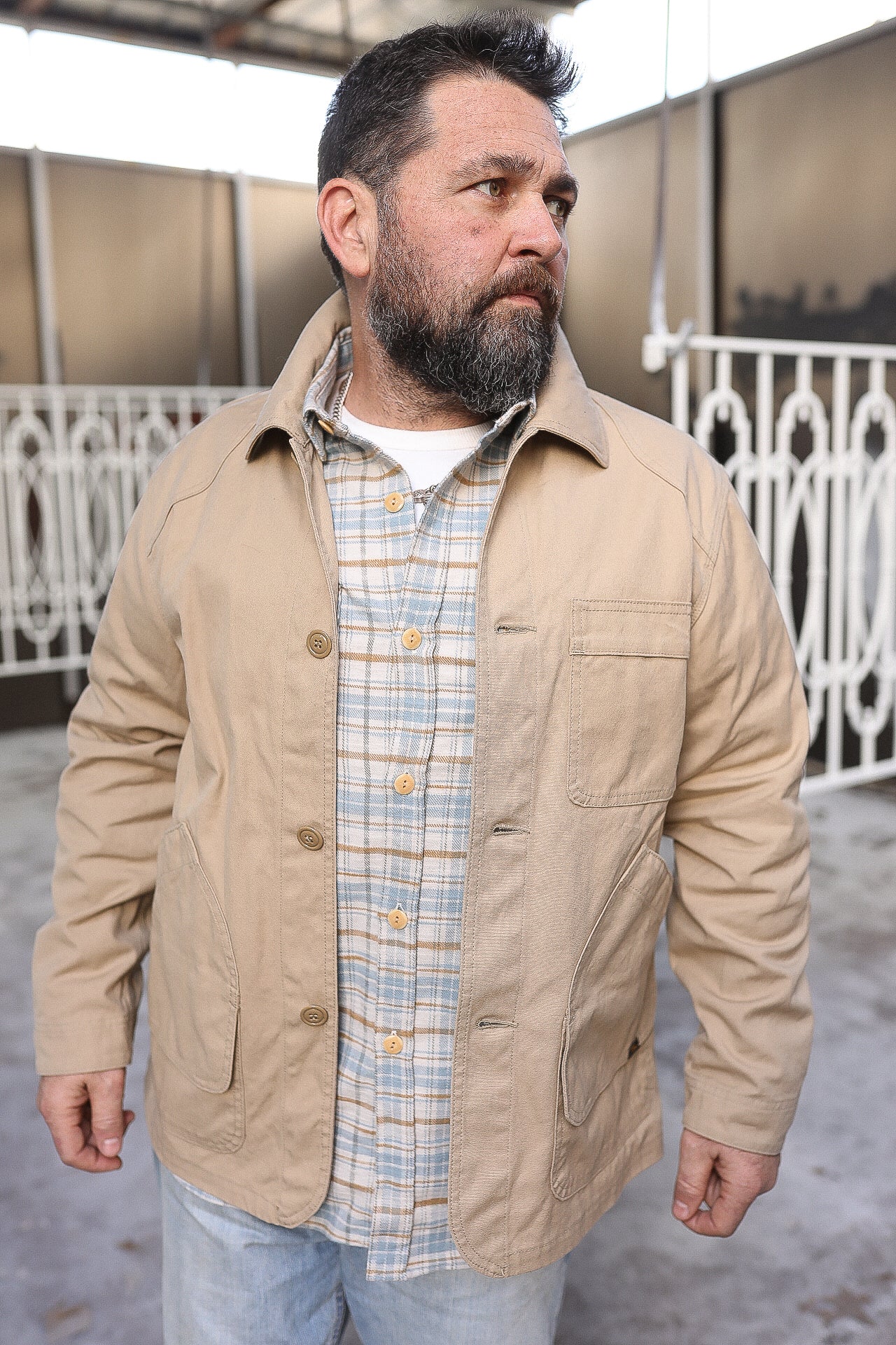 Bearded man wearing a beige work jacket over a plaid shirt outdoors by metal railing