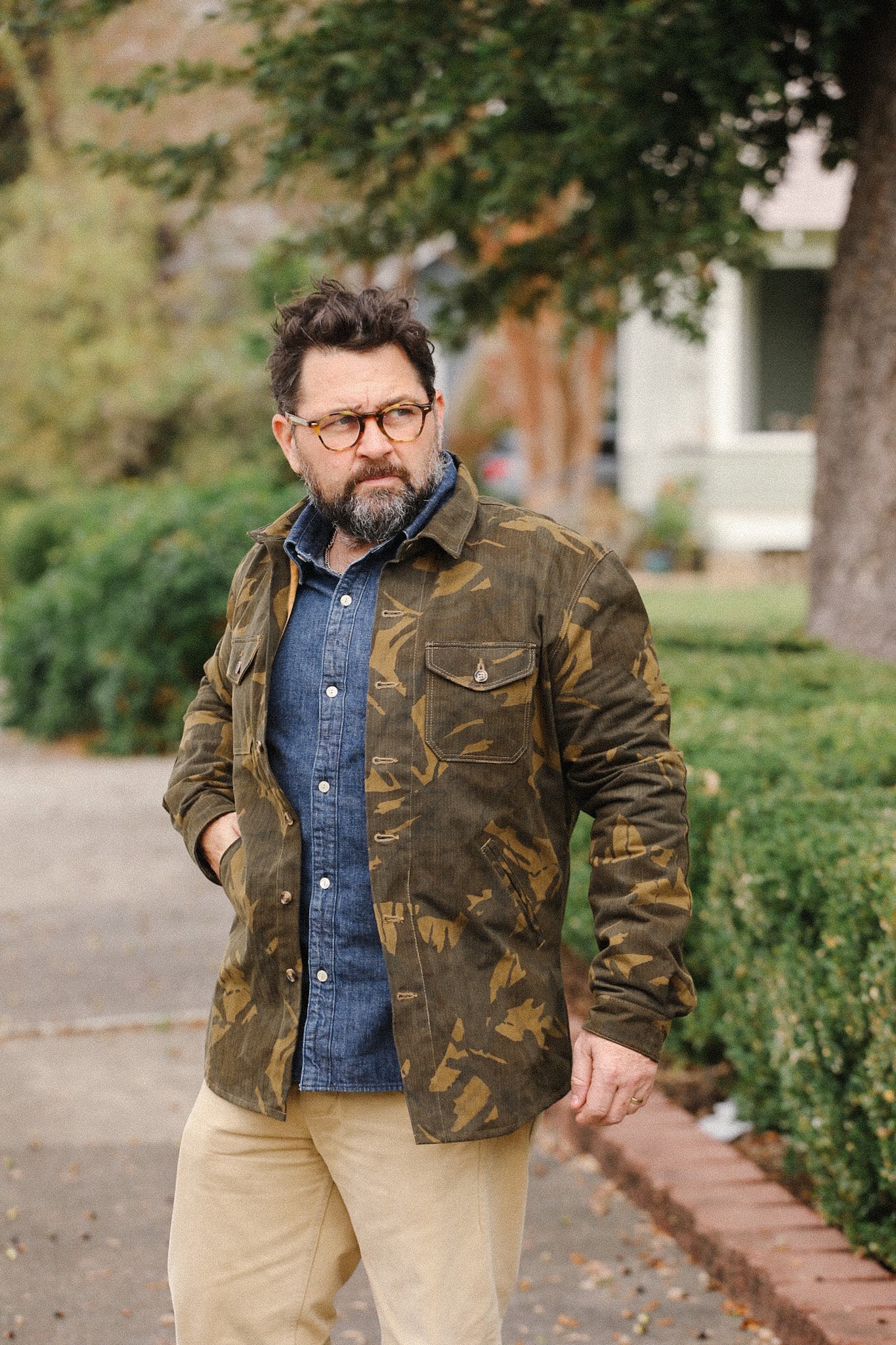 Bearded man wearing camo jacket over blue denim shirt and beige pants outdoors near bushes