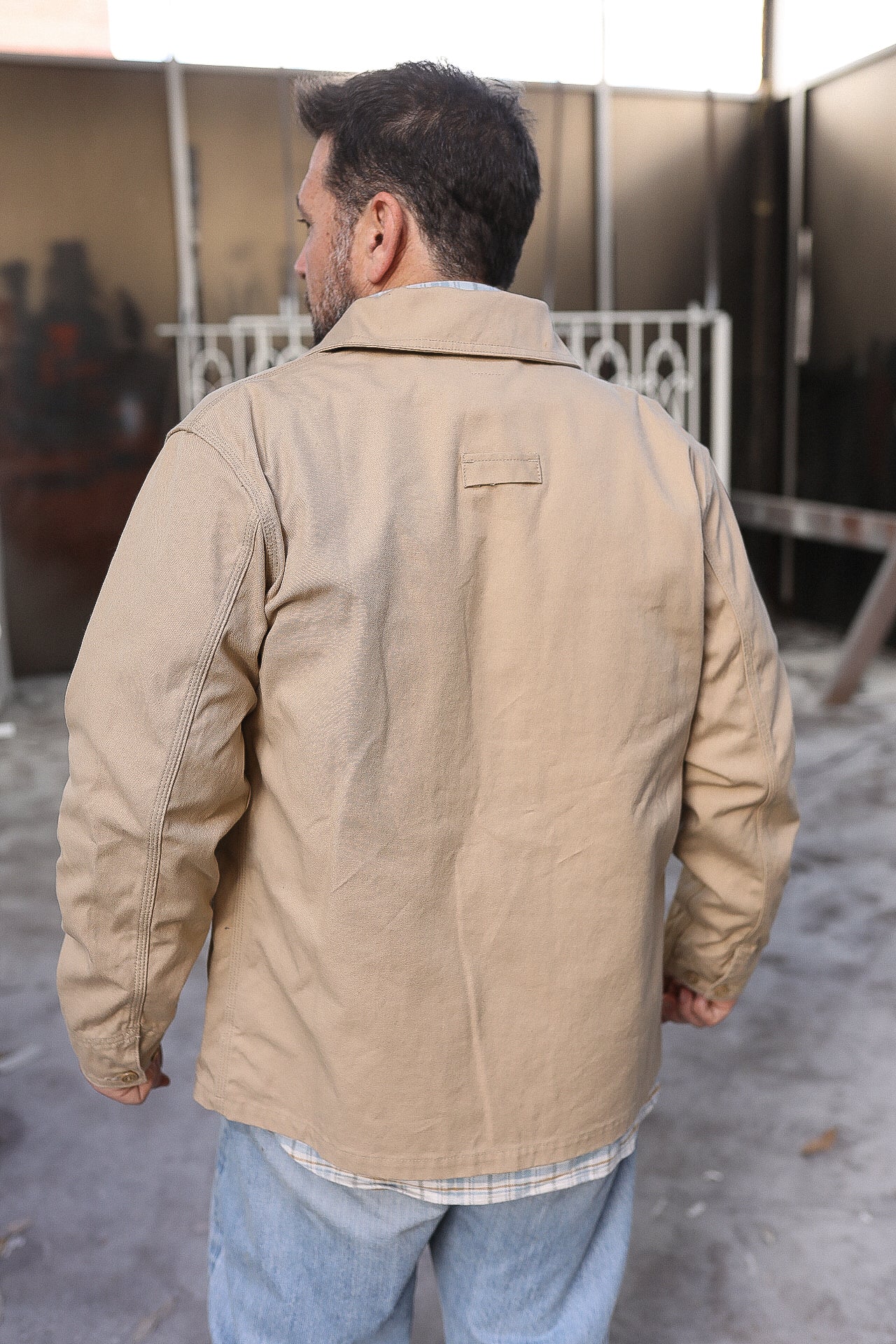 Man wearing a beige work jacket over a plaid shirt in an industrial outdoor setting