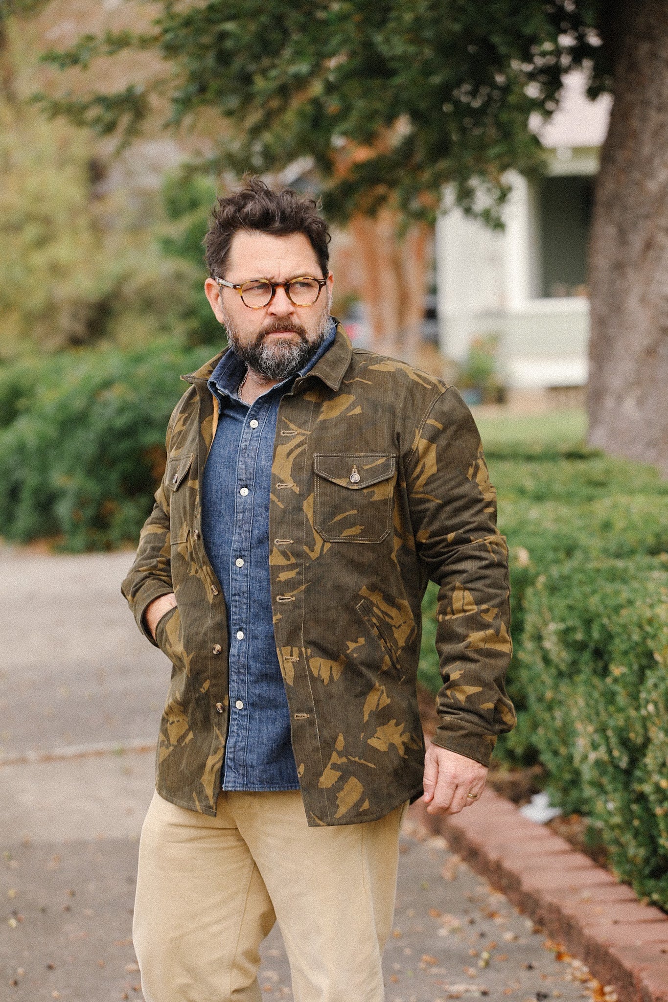 Man wearing camo jacket over blue denim shirt and beige pants outdoors by green bushes