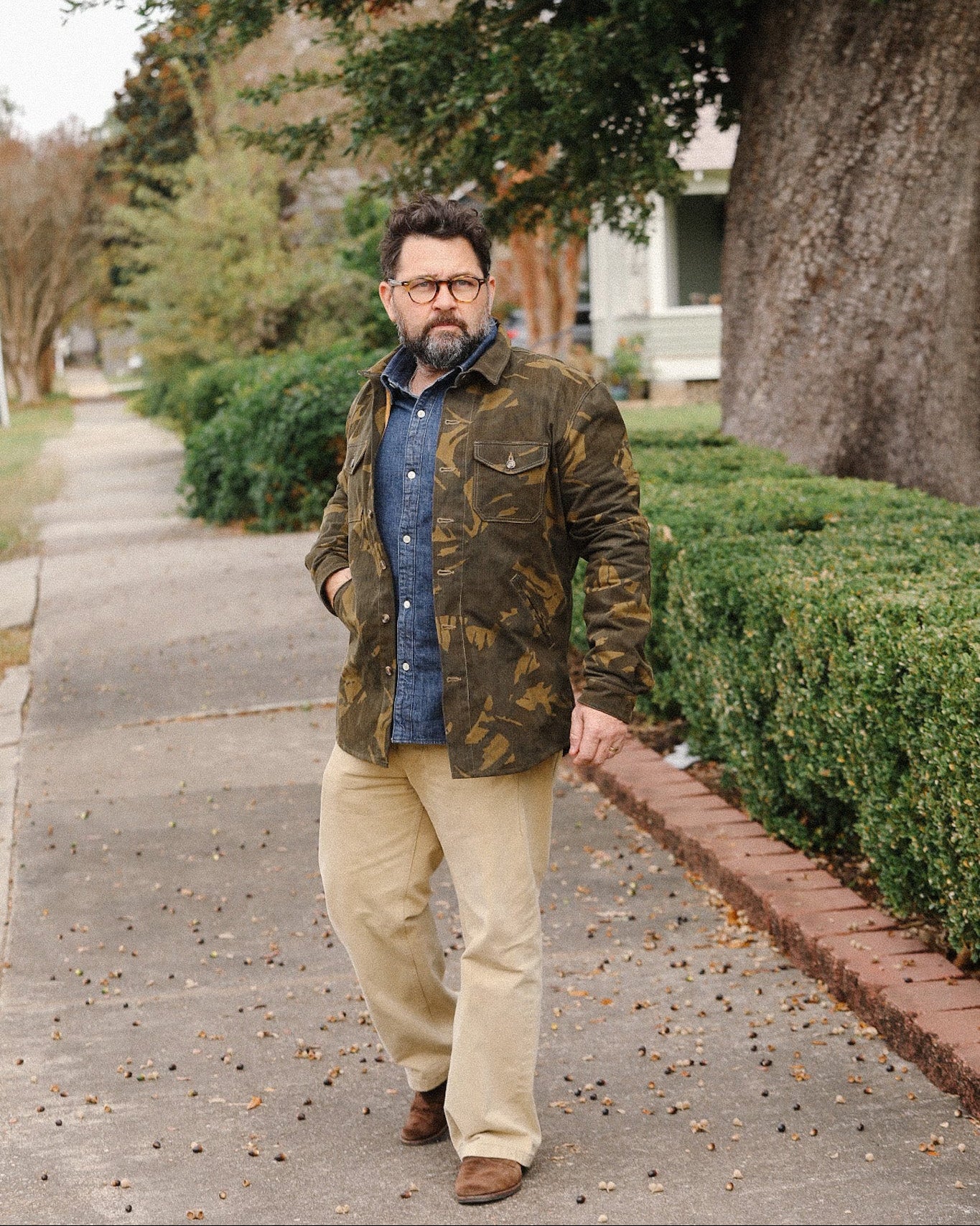 Man wearing camouflage jacket, blue denim shirt, beige pants, and brown boots walking on suburban sidewalk