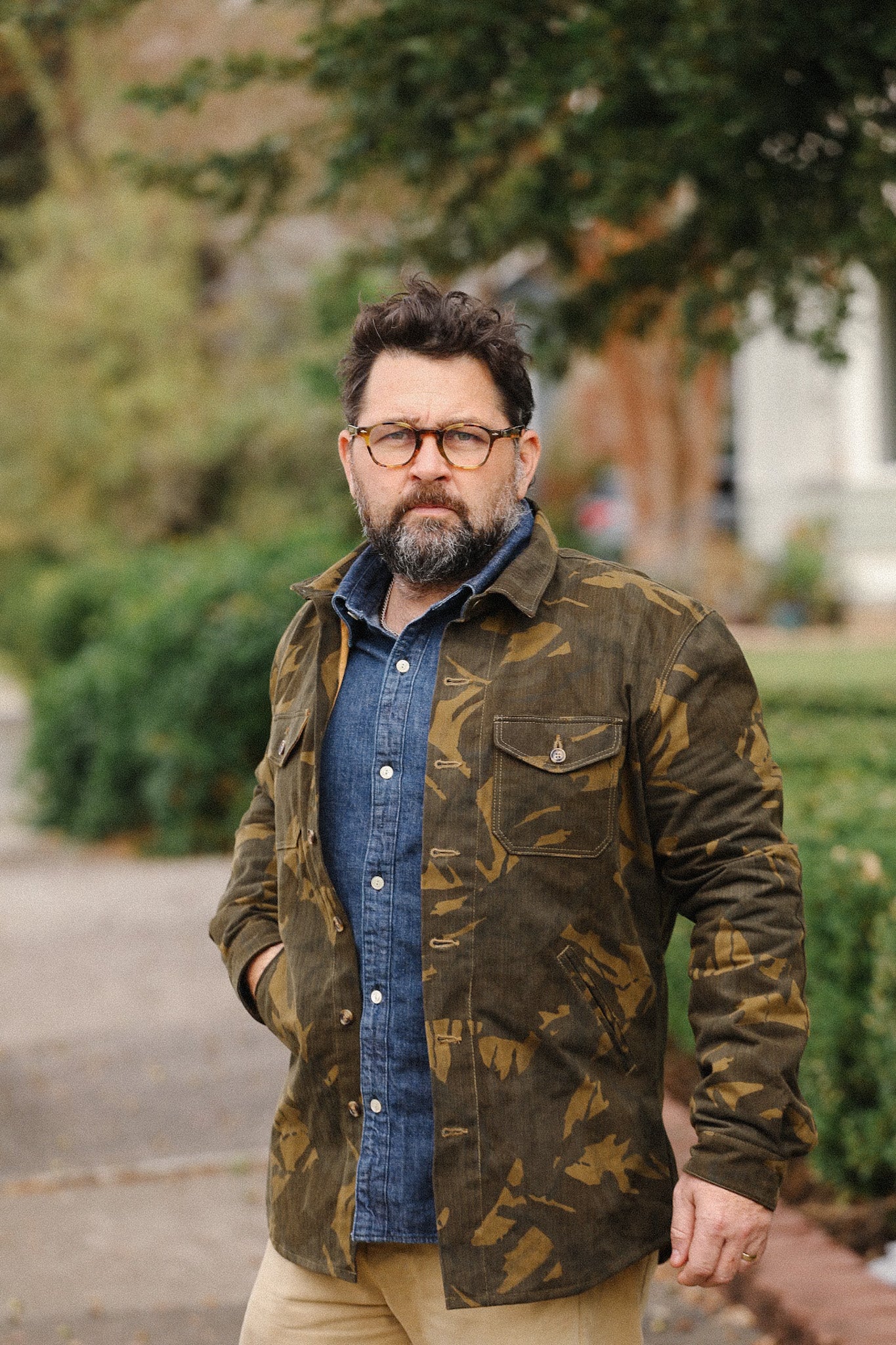 Man wearing camo jacket over denim shirt outdoors with green trees and bushes background