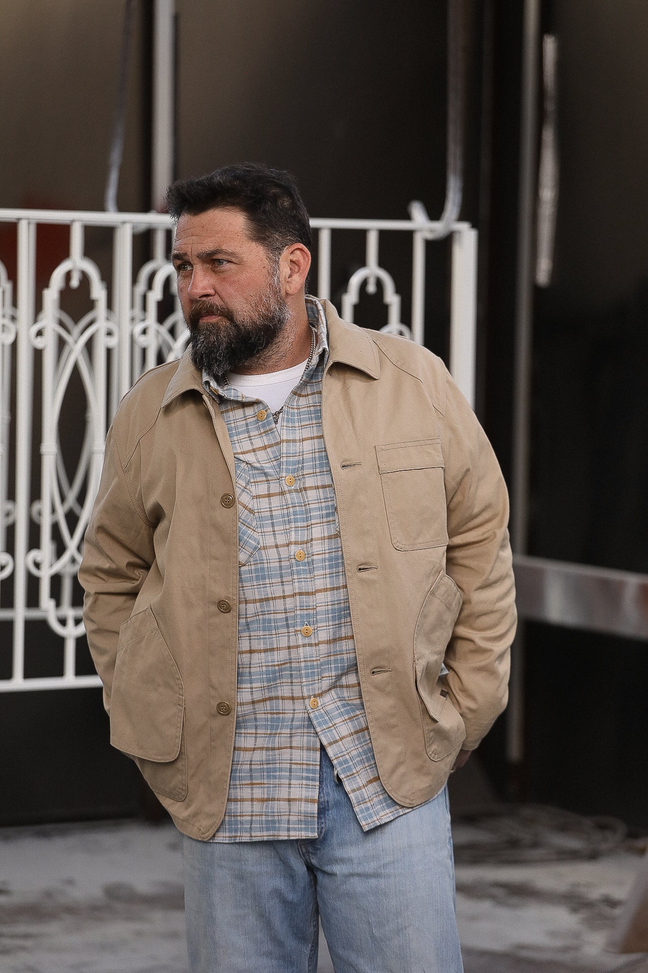 Man with beard wearing beige work jacket and plaid shirt standing outdoors near white iron gate