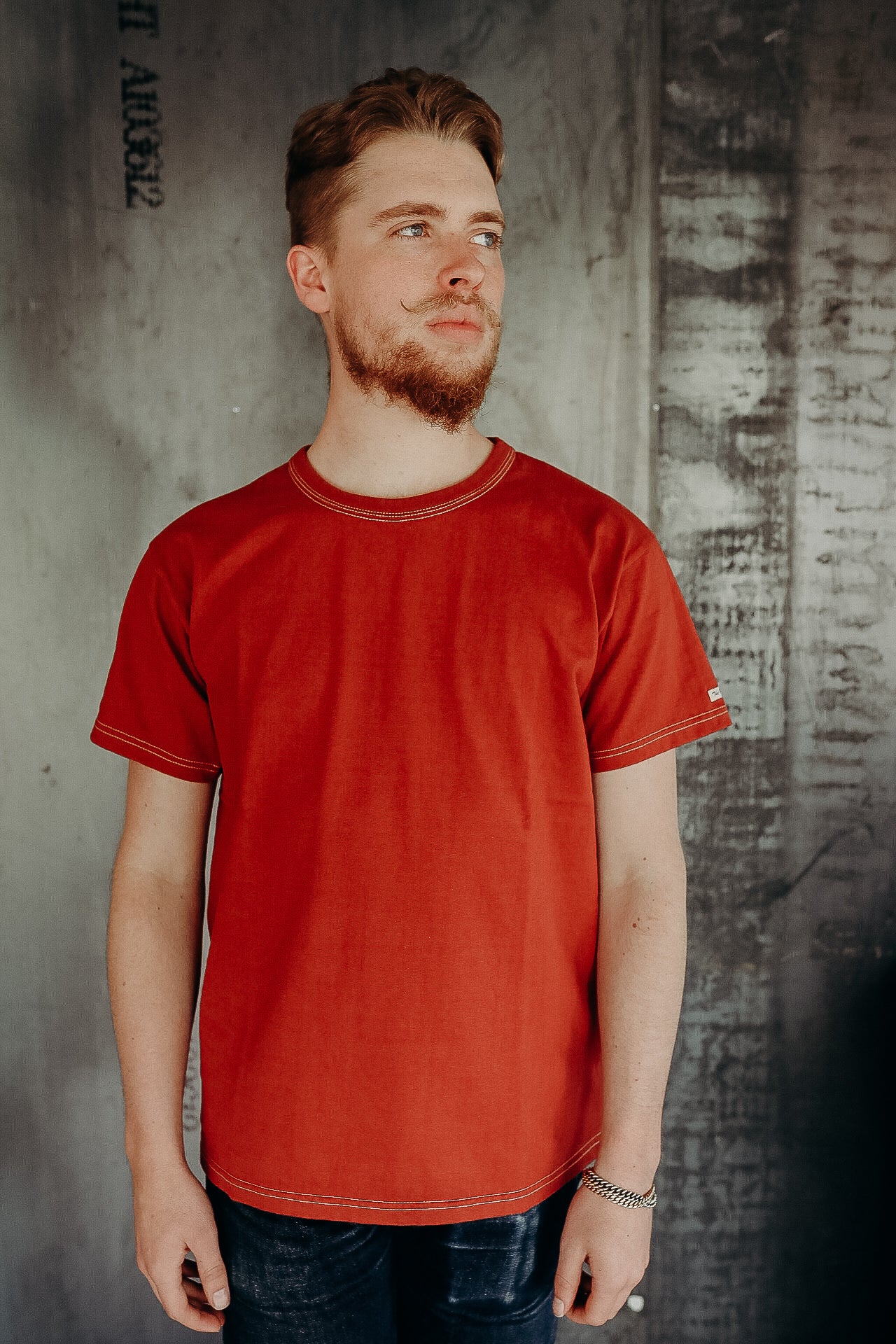 Man with styled mustache and beard wearing a red short-sleeve t-shirt against textured gray wall