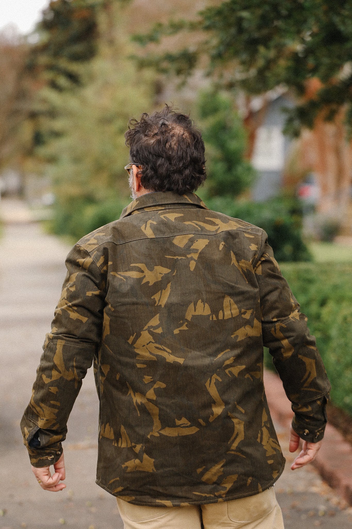 Man wearing camo patterned jacket walking outdoors on a tree-lined sidewalk