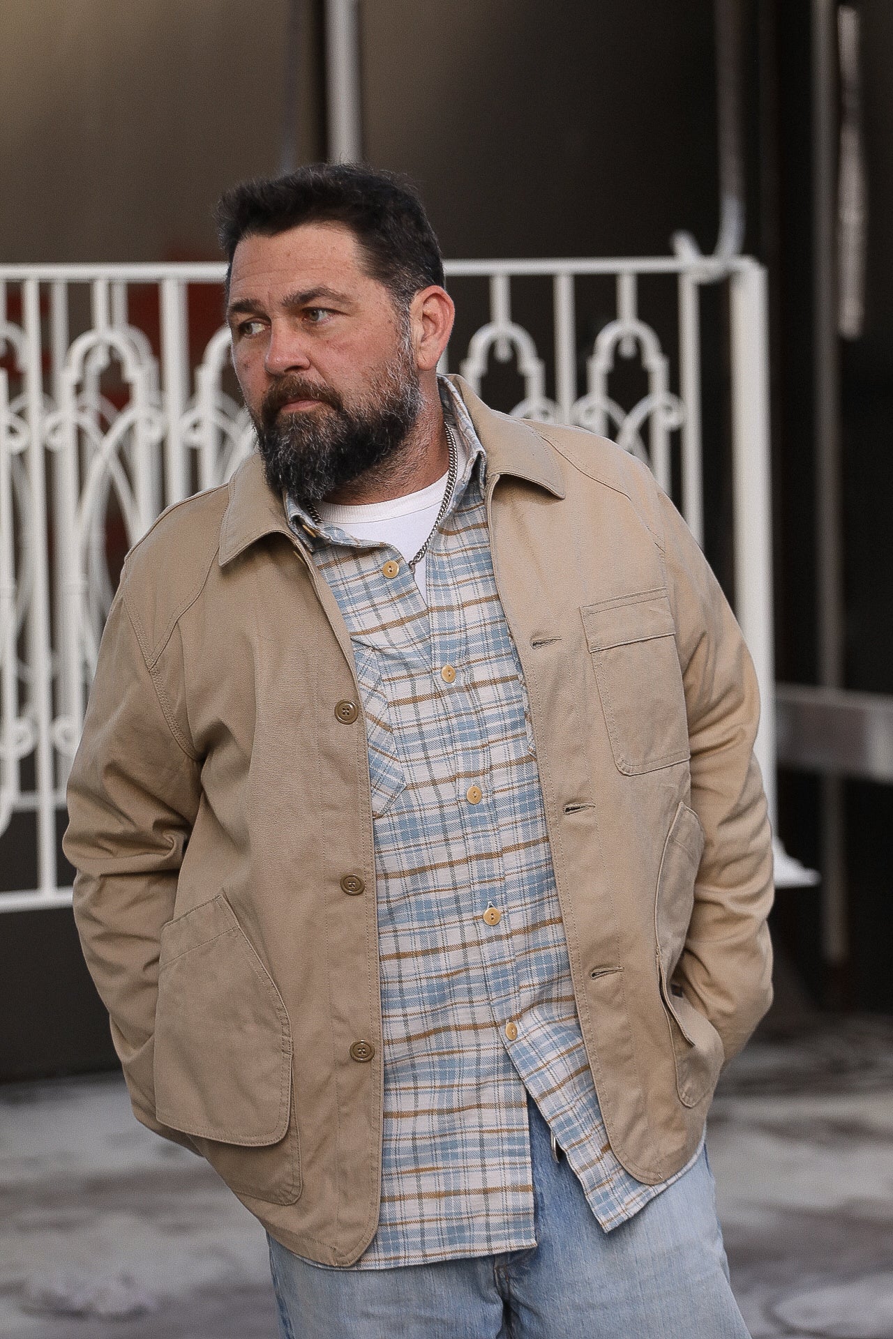Man wearing beige work jacket and plaid shirt with blue jeans standing outdoors by white metal fence