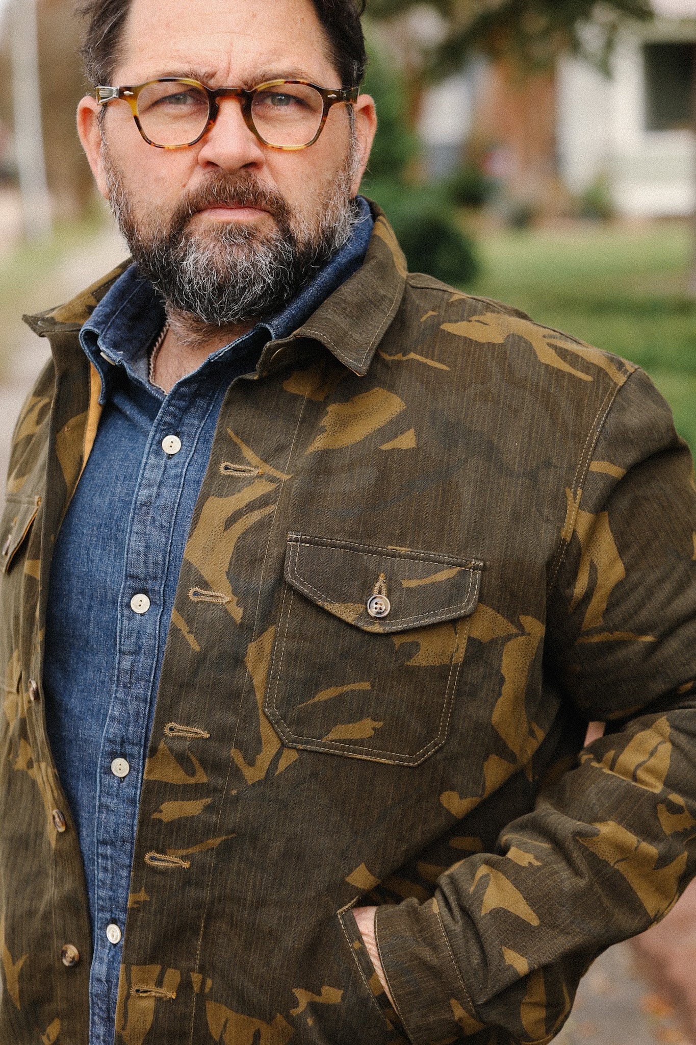 Bearded man wearing tortoiseshell glasses, camouflage jacket, and denim shirt outdoors