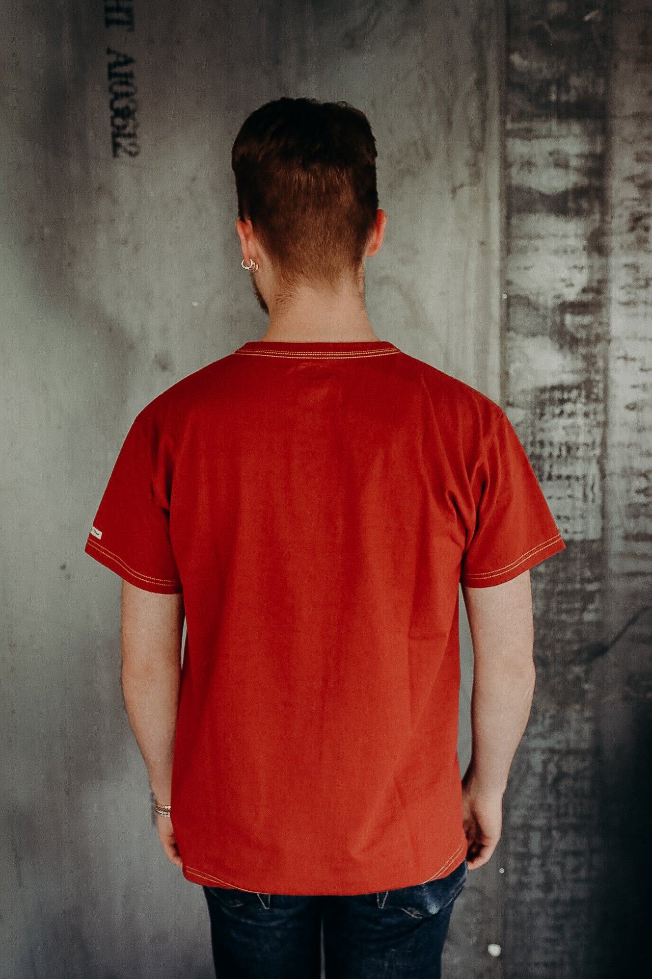 Man wearing a rust red short-sleeve shirt with visible stitching, standing against a textured industrial wall