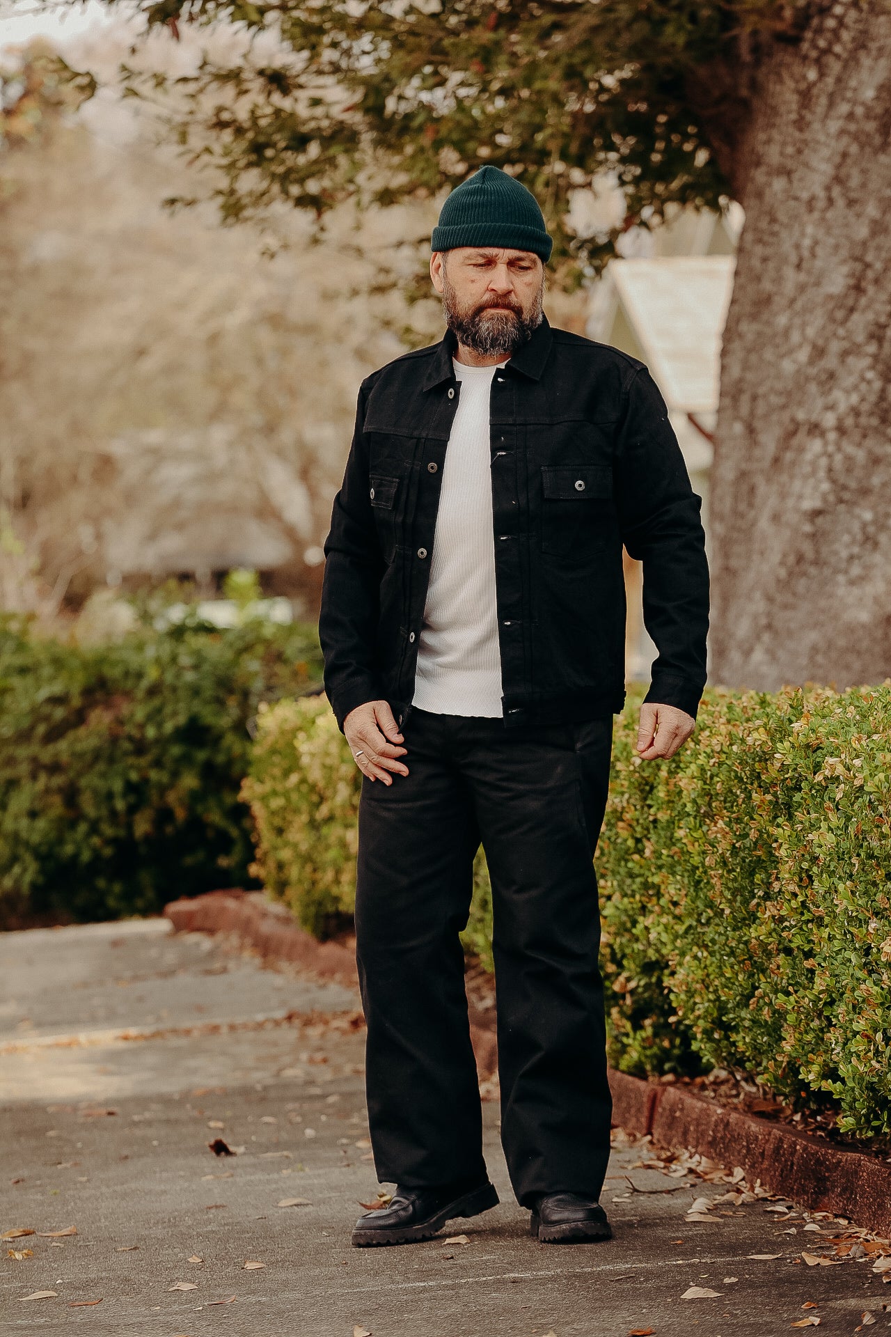 Man wearing black denim jacket, black pants, green beanie, and boots standing outdoors near bushes