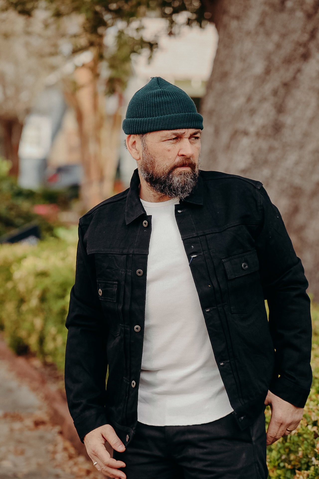 Man wearing black denim jacket and dark green beanie outdoors with trees and bushes