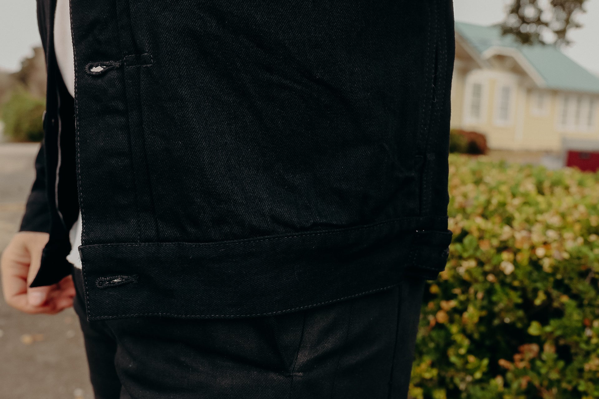 Close-up of a person wearing a black denim jacket outdoors with greenery and houses in the background