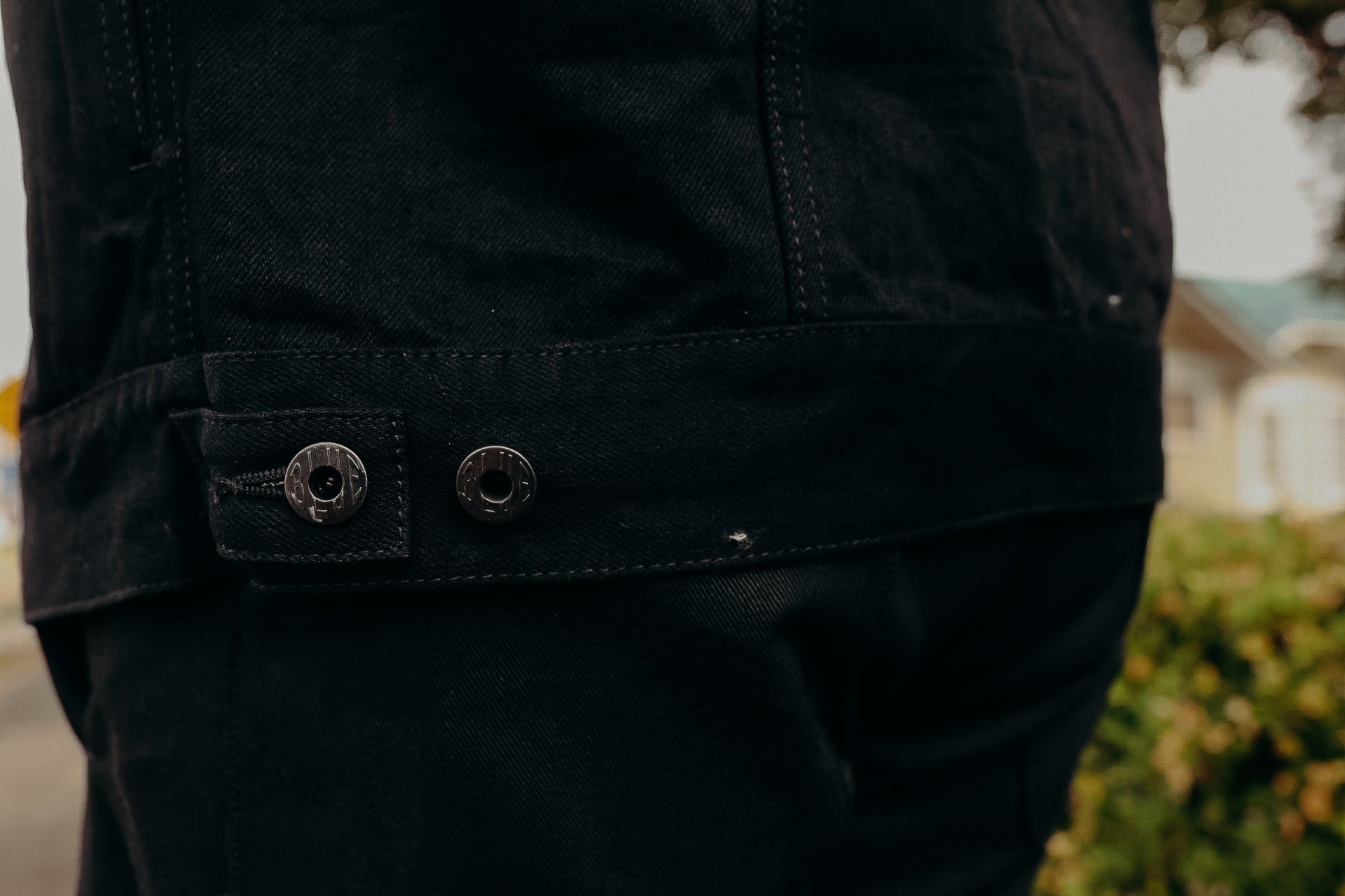 Close-up of a black denim jacket hem with two metal buttons engraved with 'IRON SHOP' outdoors