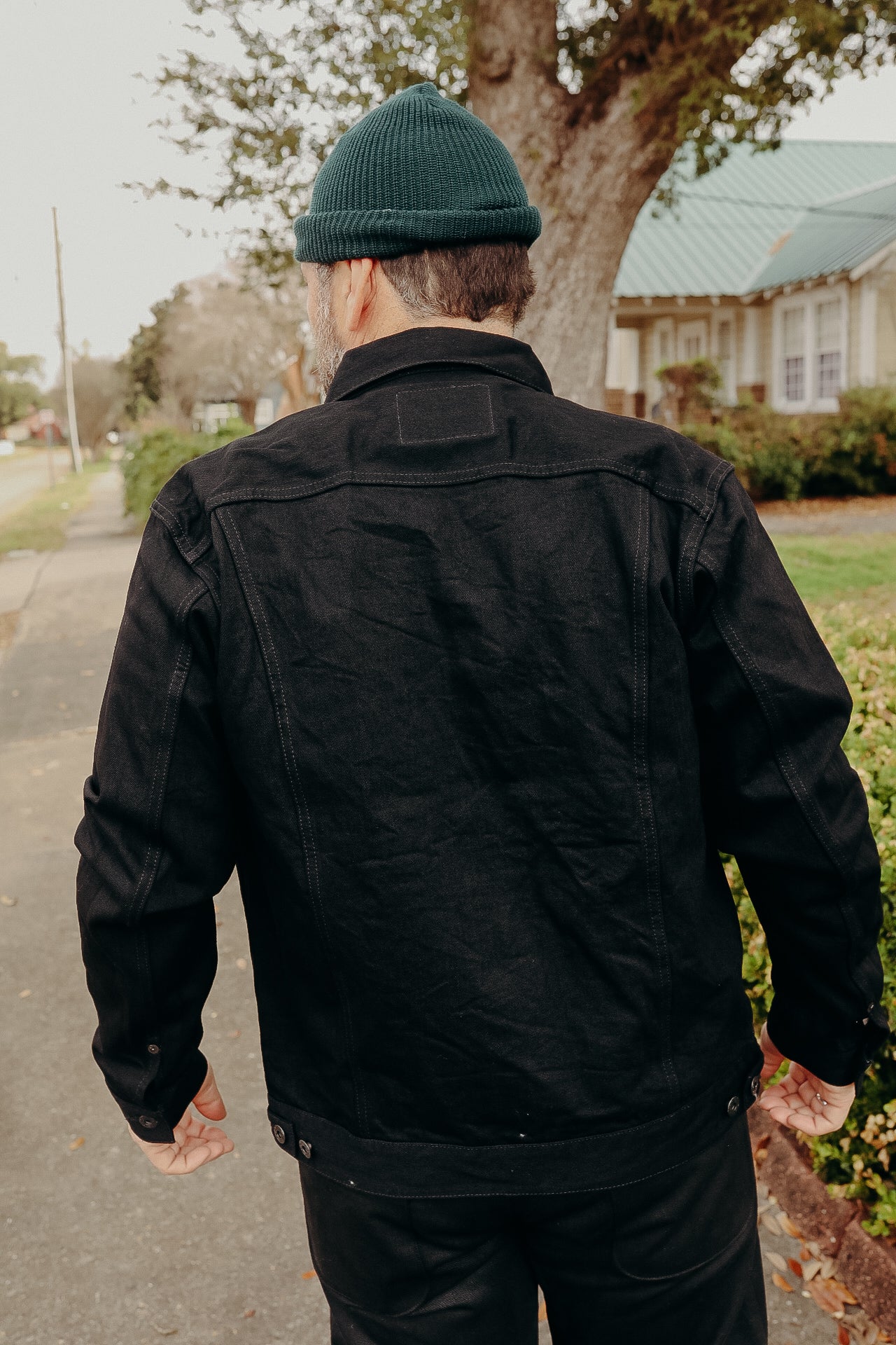 Man wearing black denim jacket and green knit beanie walking on suburban sidewalk near trees and houses