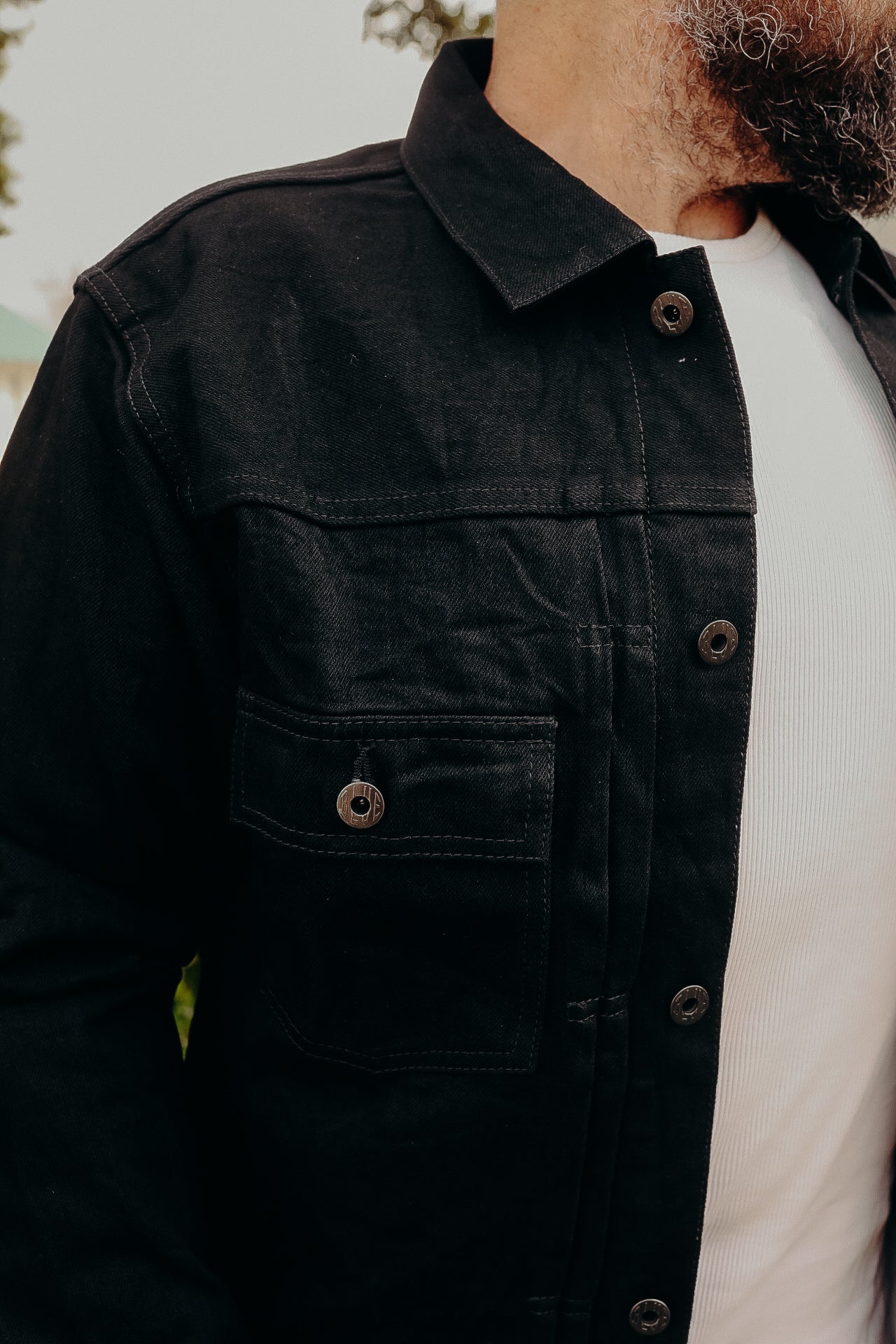 Close-up of a man wearing a black work jacket with metal buttons over a white ribbed shirt