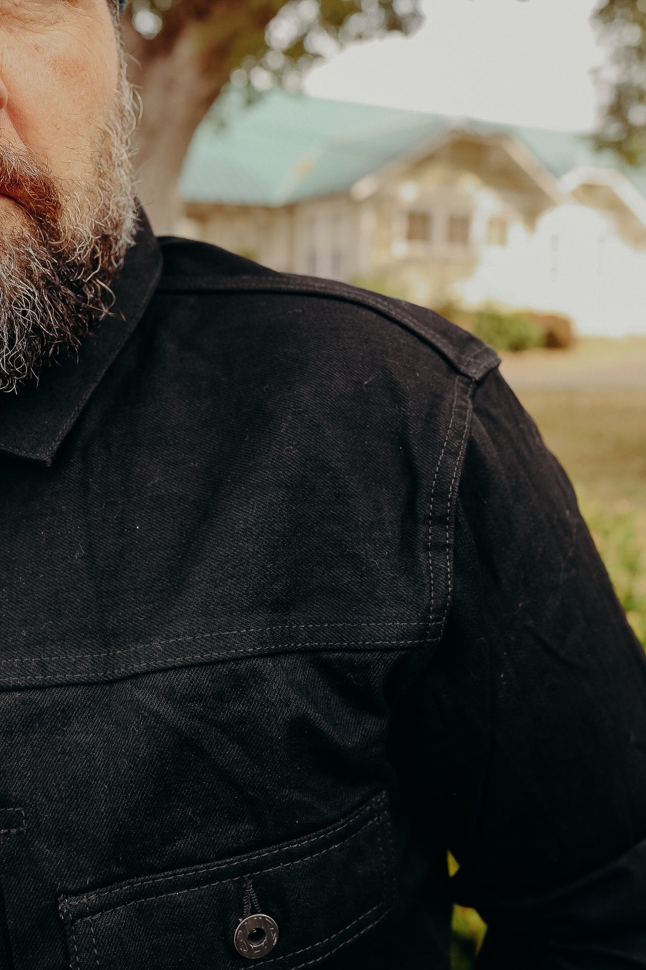 Close-up of man's beard and black denim jacket with buttoned pocket, blurred house in background